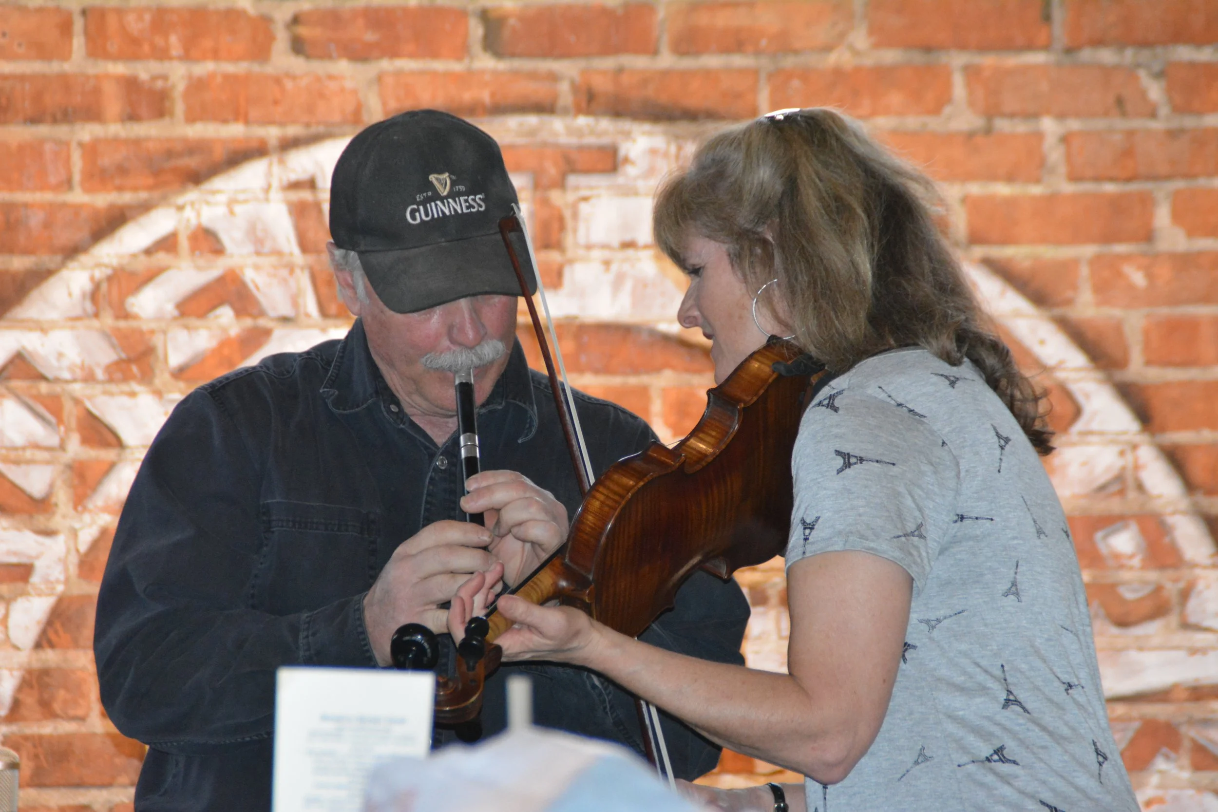An older man playing a flute and a woman playing a violin, standing close together against a brick wall background.