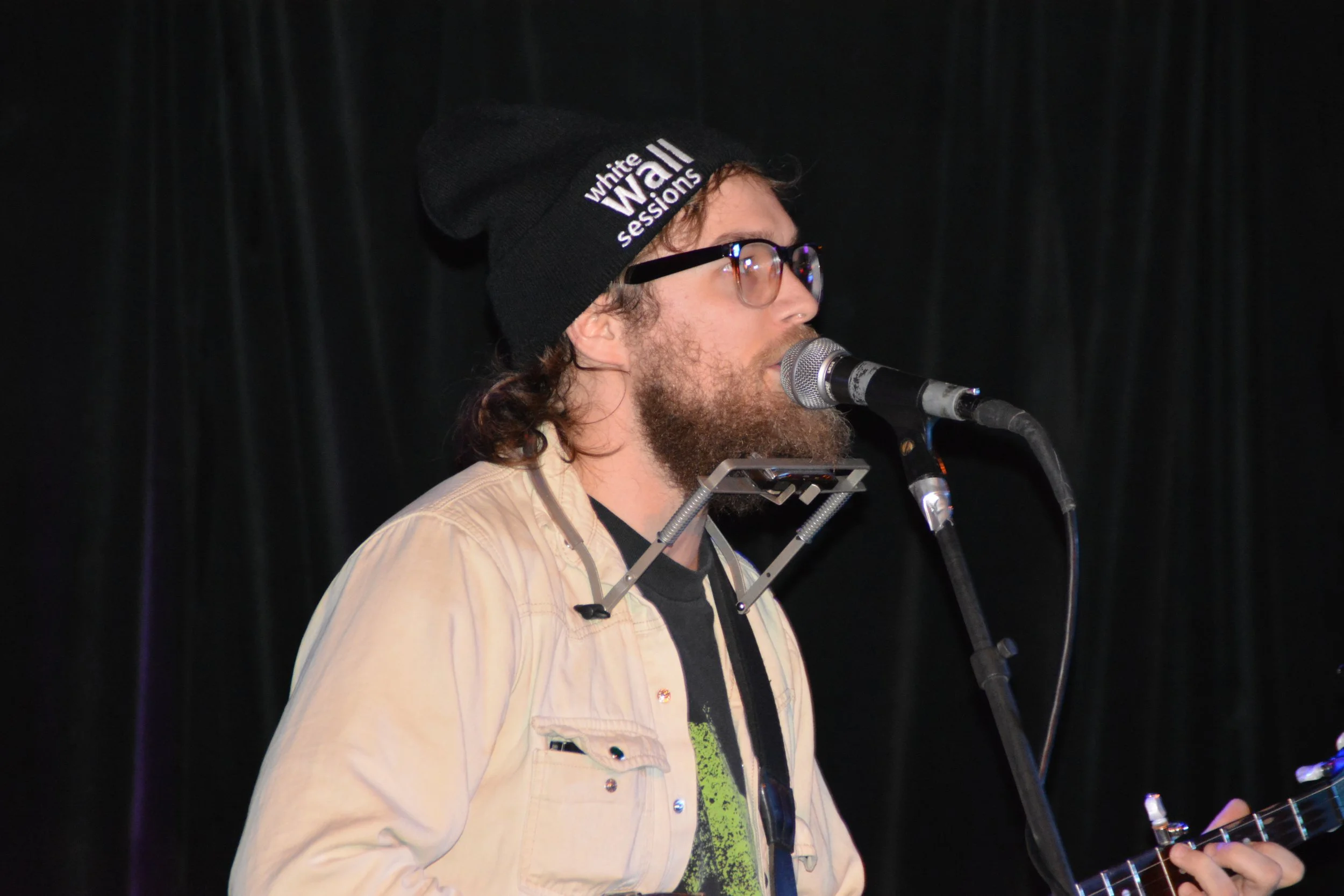 A man with curly hair and a beard wearing glasses, a black beanie that says 'white Wall sessions', a beige jacket, and a black t-shirt, singing into a microphone and holding a guitar on a dark stage.