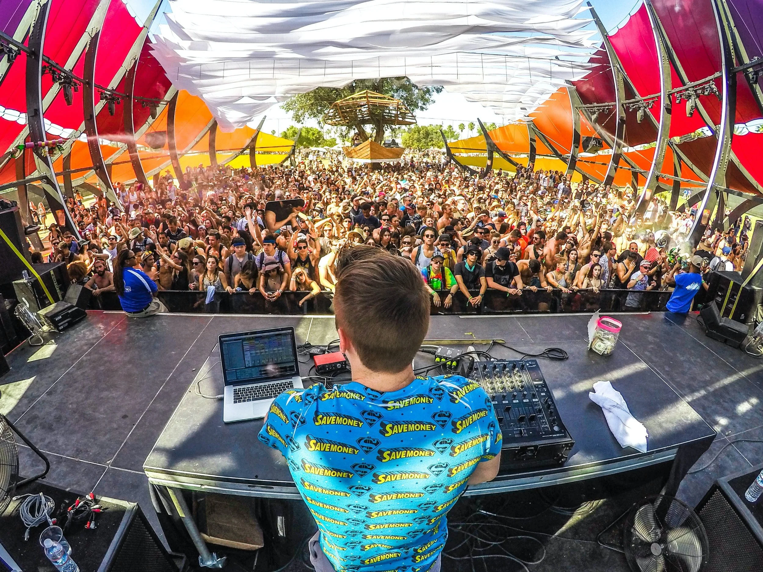 DJ performing at an outdoor music festival with a large crowd dancing under colorful canopy structures.