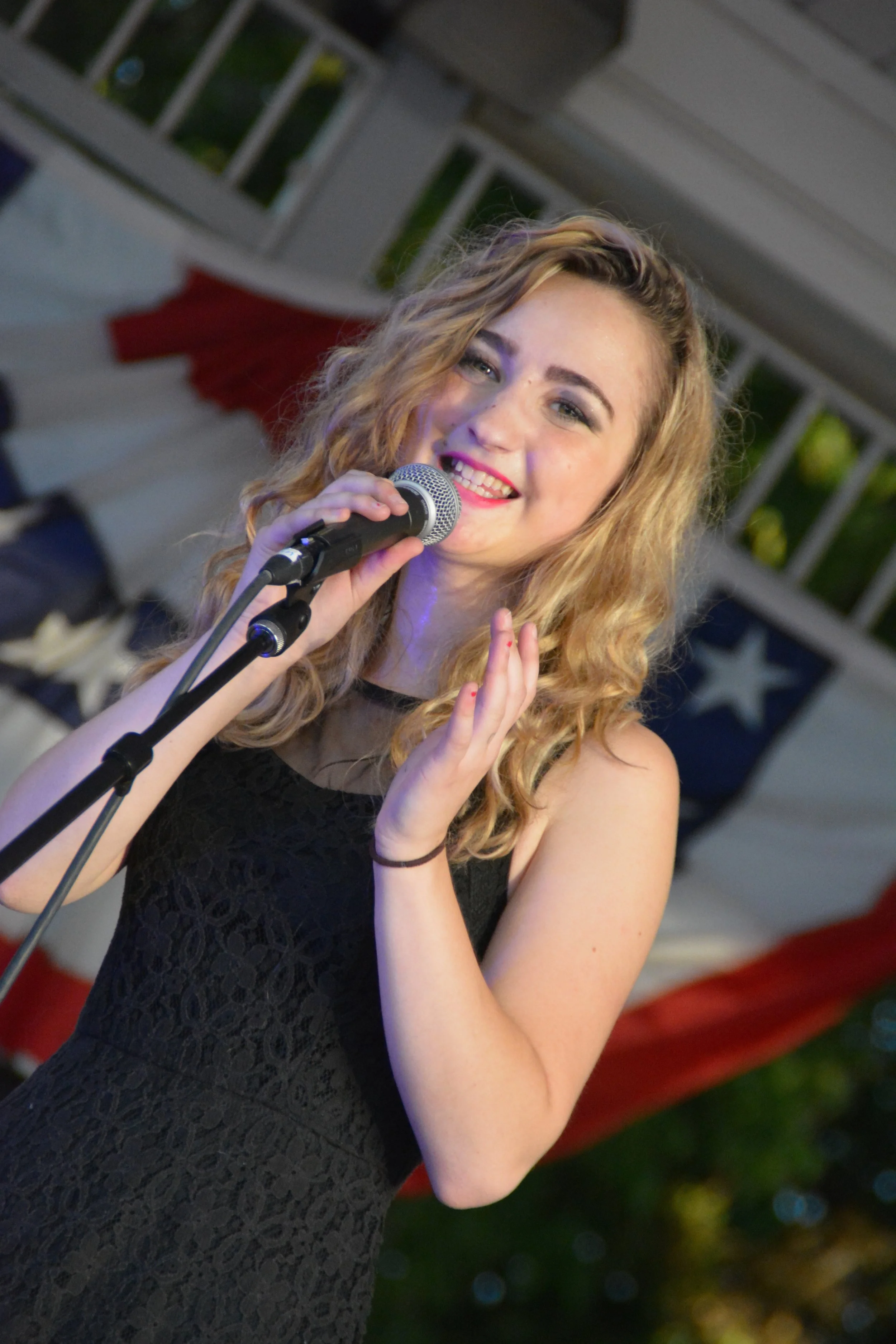 A young woman with curly blonde hair singing or speaking into a microphone at an outdoor event, with patriotic decorations and a porch railing in the background.