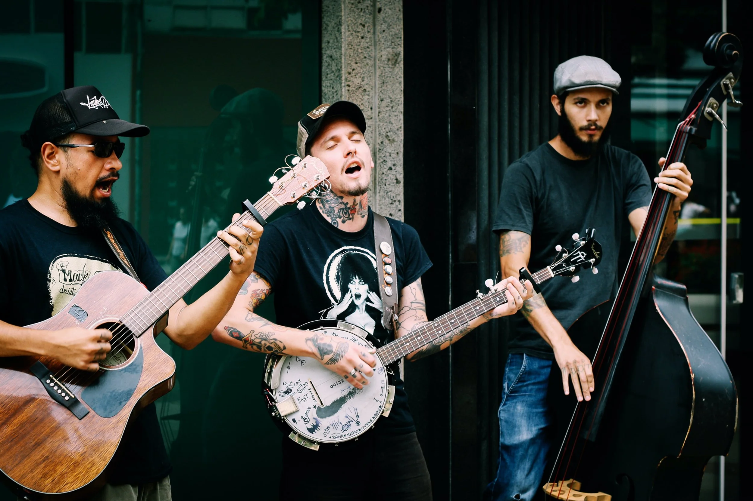 Three male street musicians performing with guitars and a double bass in an urban setting.