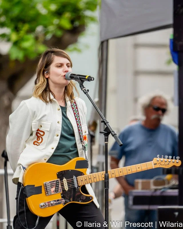 Young woman singing into a microphone while holding an electric guitar at an outdoor event, with a man in sunglasses and a blue shirt in the background.