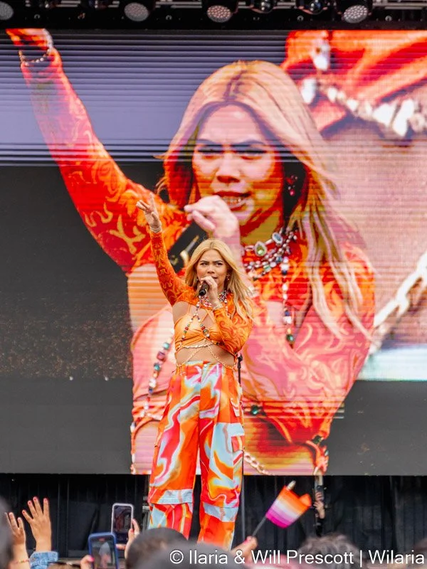 Performer on stage singing into a microphone with a large screen behind her displaying her image, wearing an orange and red patterned outfit, with hands raised and an audience waving a small flag.