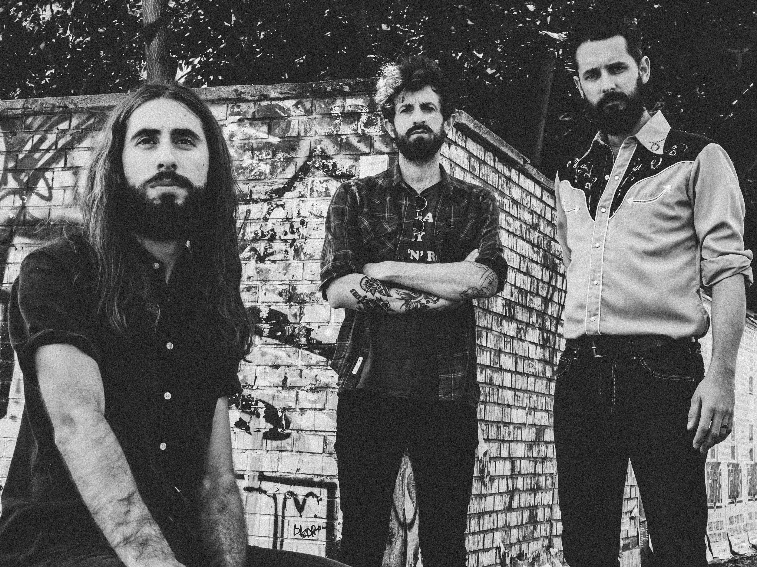 Black and white photo of three men standing in front of a brick wall with graffiti, looking at the camera with serious expressions.