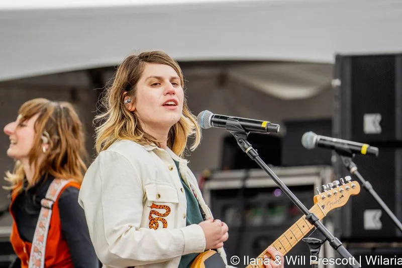 Two women performing on stage with microphones and musical instruments at an outdoor event.