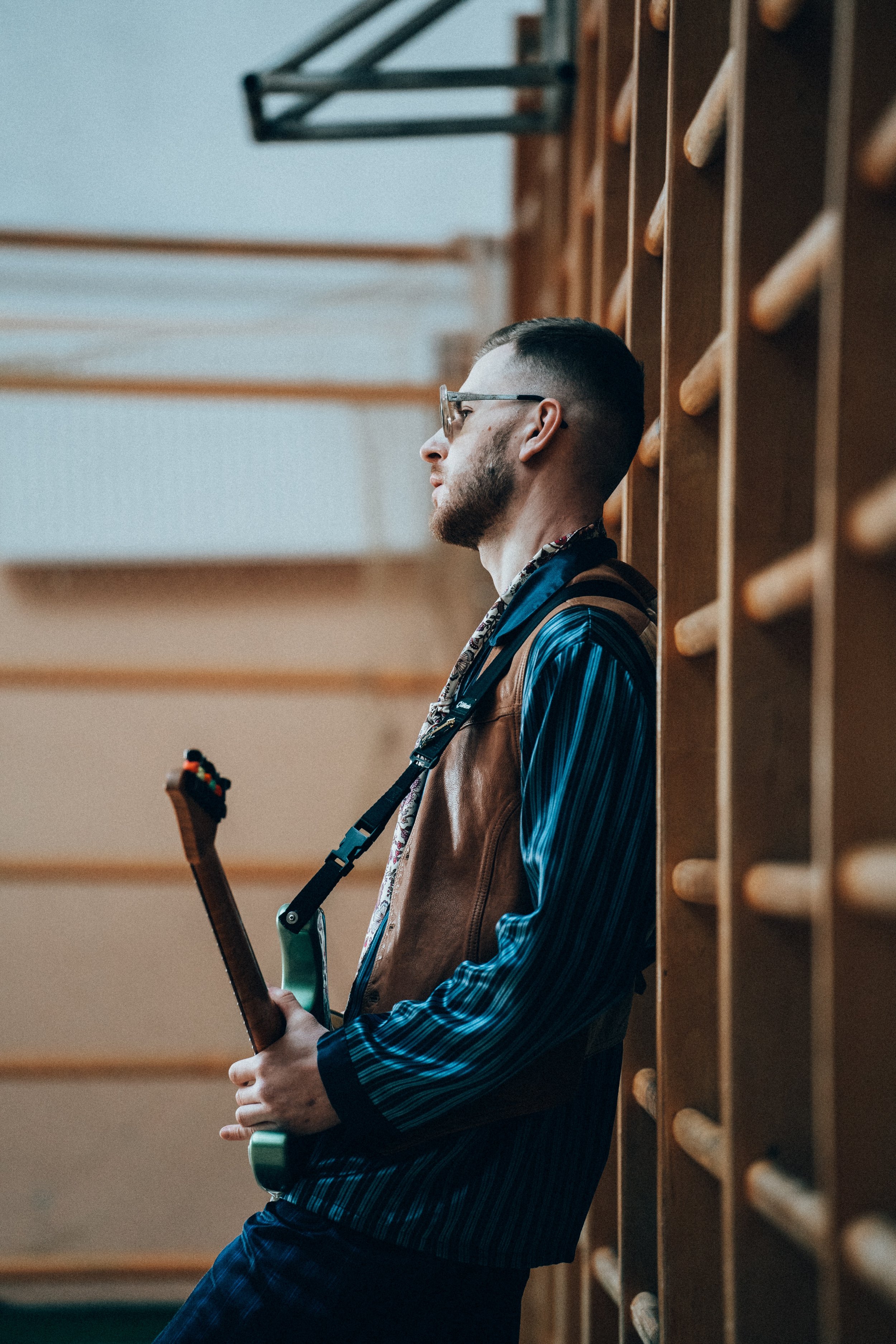 A man with glasses and a beard leaning against a wooden wall with horizontal rods, holding an electric guitar, in what appears to be a music practice room or studio.