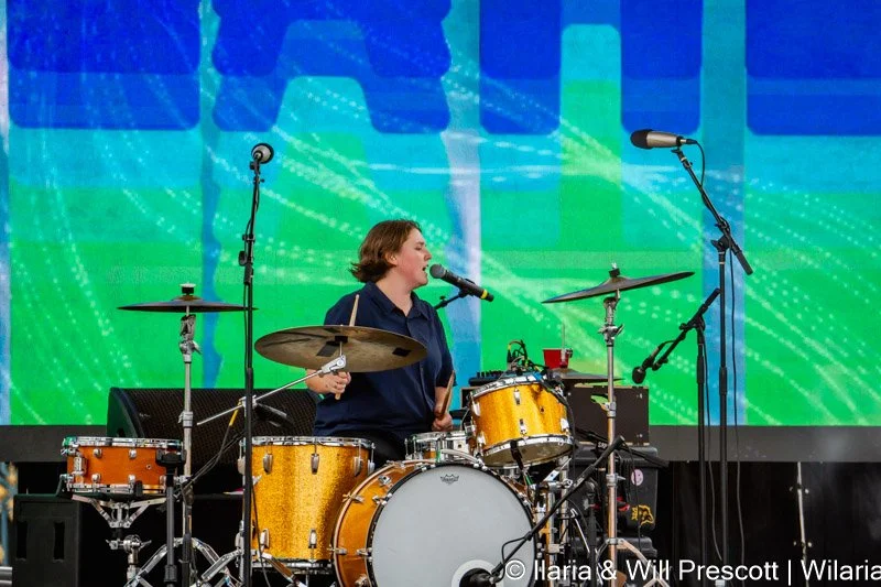 A woman singing into a microphone while playing a drum set on a stage with a vibrant digital backdrop.