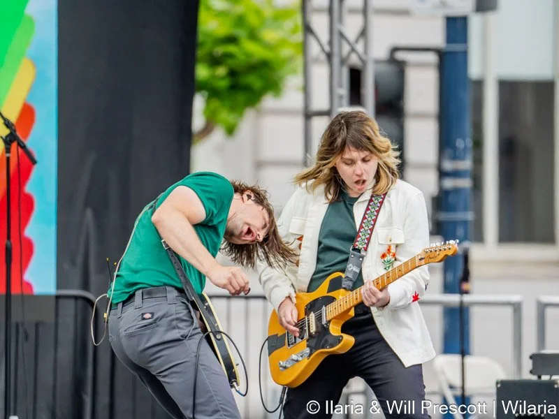 Two young women playing electric guitars passionately on an outdoor stage, with one wearing a green shirt and the other a white jacket. They appear to be performing intensely.