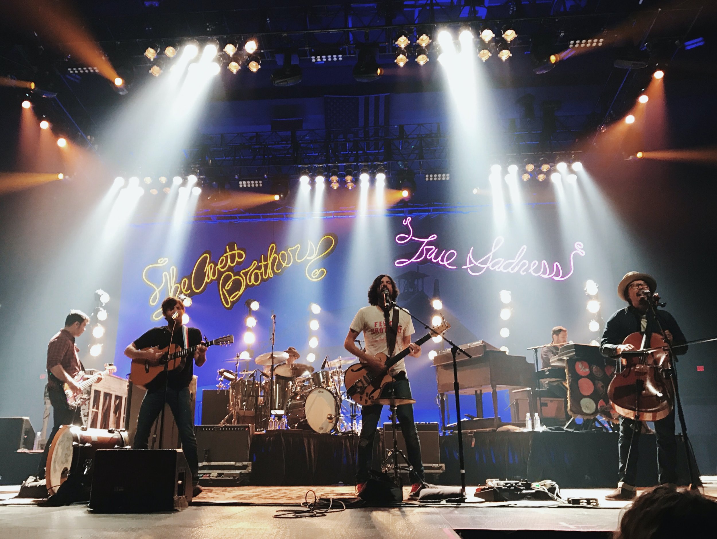 A band performing live on stage with five members, lights, and neon signs in the background reading 'She Acted Like a Brother' and 'True Sadness'.