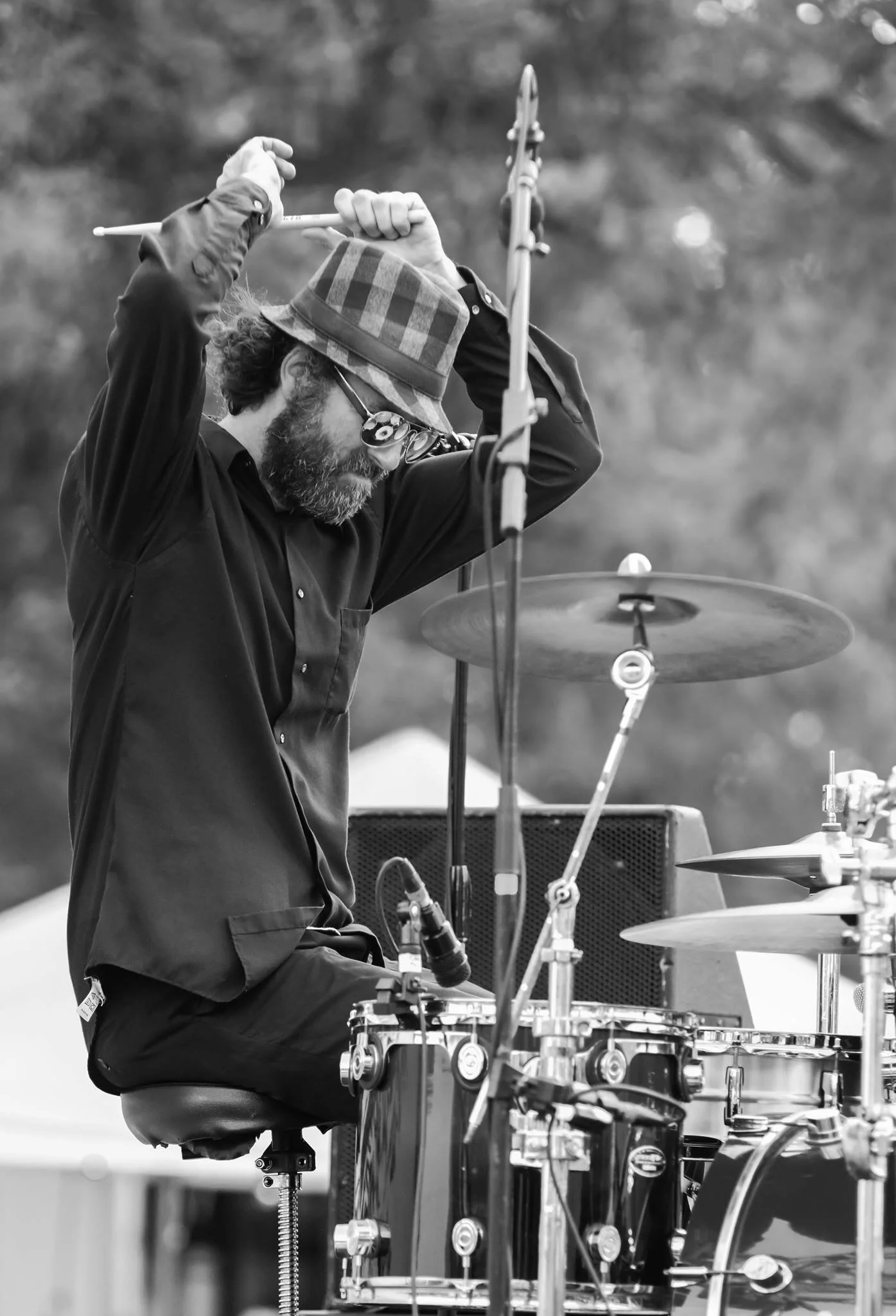 A man plays drums outdoors, wearing a plaid hat, glasses, and a black shirt, captured in black and white.