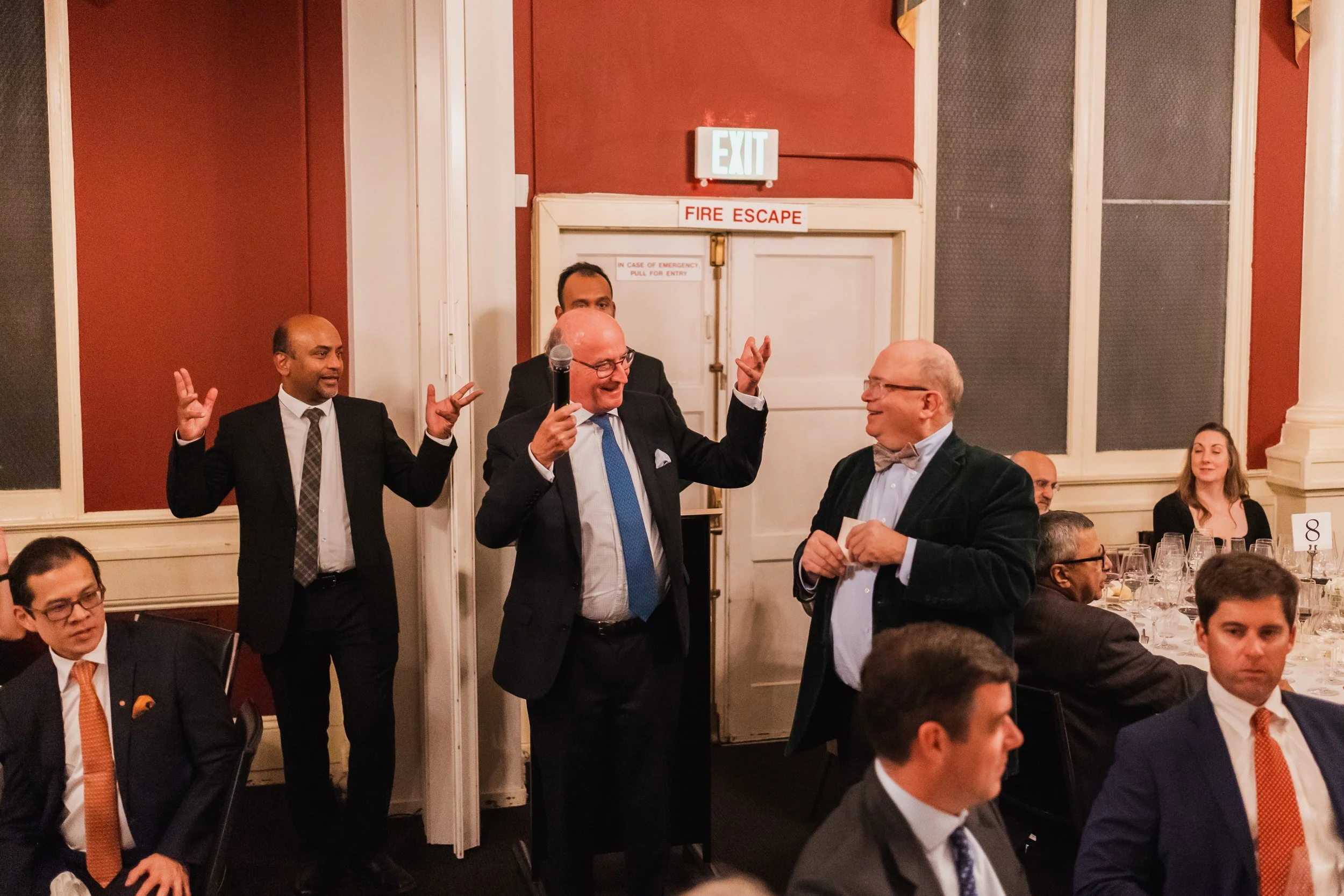 A group of men in formal attire are gathered in a banquet hall. One man with glasses and a bow tie is holding a card, smiling, as another man with glasses and a suit speaks. Two men in the background are gesturing with their hands; all are engaged in
