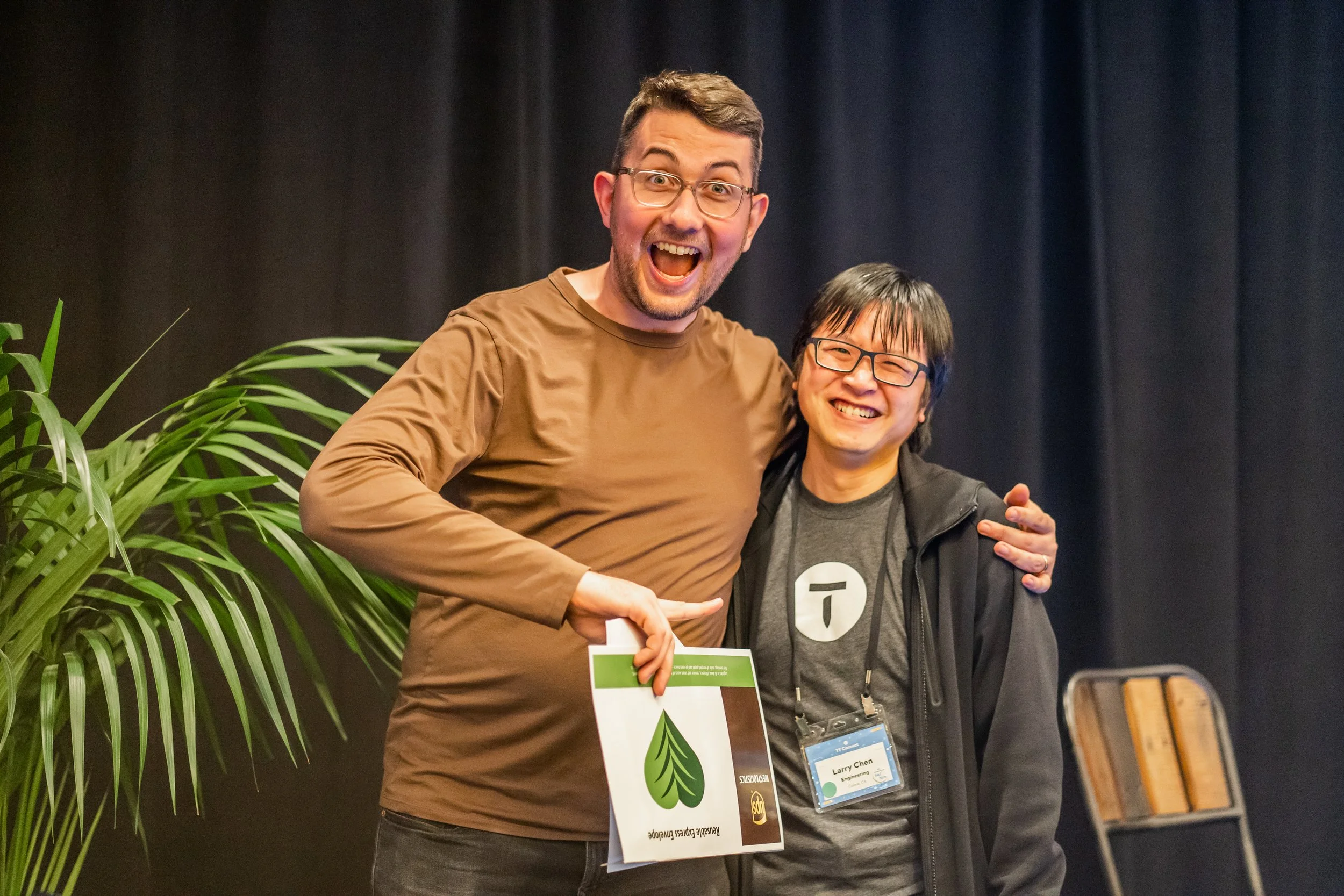 Two men smiling happily and posing for a photo at an event. One man is holding a sign that says 'Sustainable Pressures' with a leaf logo. The background is dark with a large plant on the left and a chair on the right.