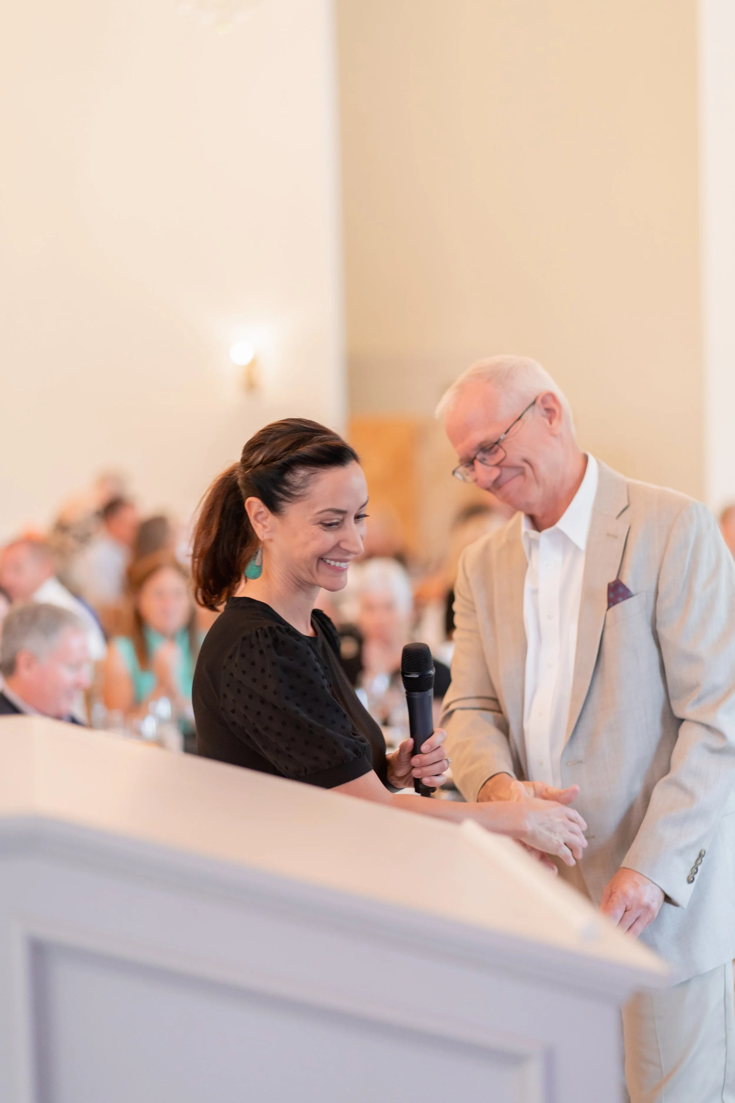 A woman with dark hair holding a microphone smiles and holds hands with an older man wearing glasses and a beige suit at a formal event with many seated guests in the background.