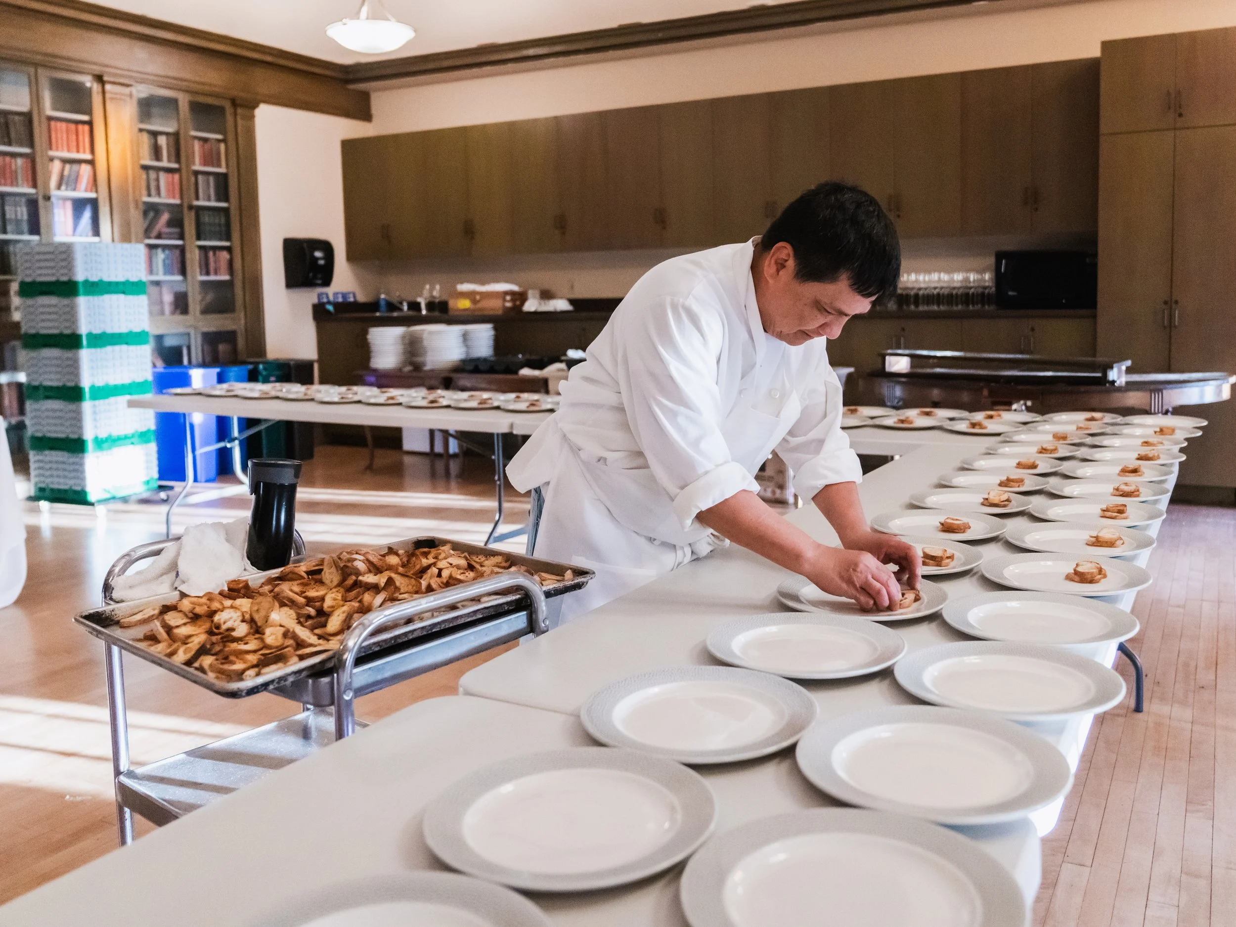 A chef preparing dishes in a restaurant kitchen, with rows of empty white plates and some cooked chicken on a tray.