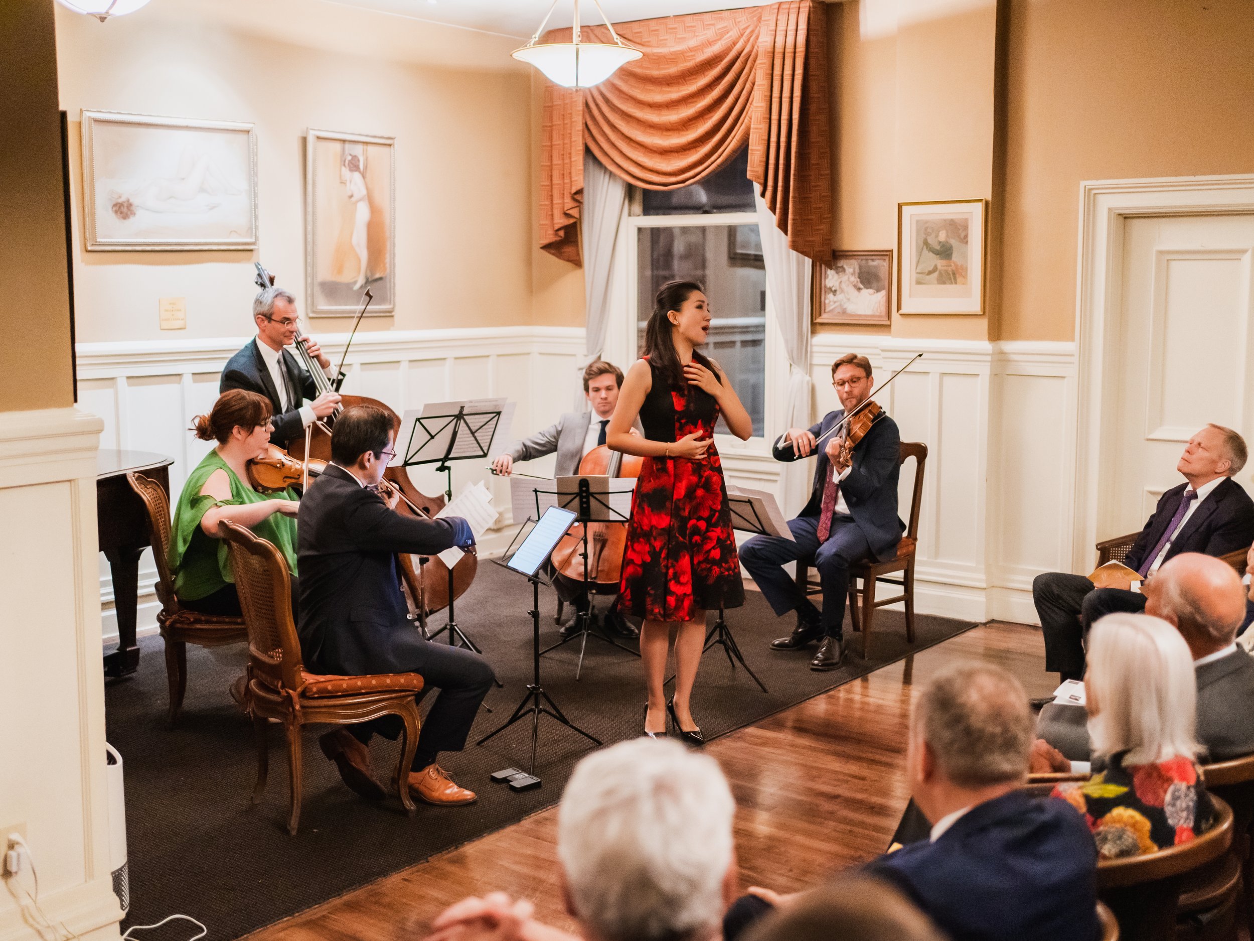 A woman in a red and black dress singing in front of a small orchestra during a performance in an elegant room with trimmed walls, framed artwork, and a large window with draped curtains.