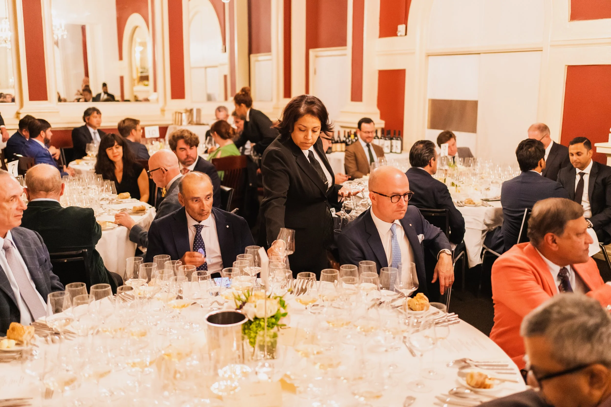 People in formal attire seated at dinner tables in a banquet hall, with a waitress serving wine.