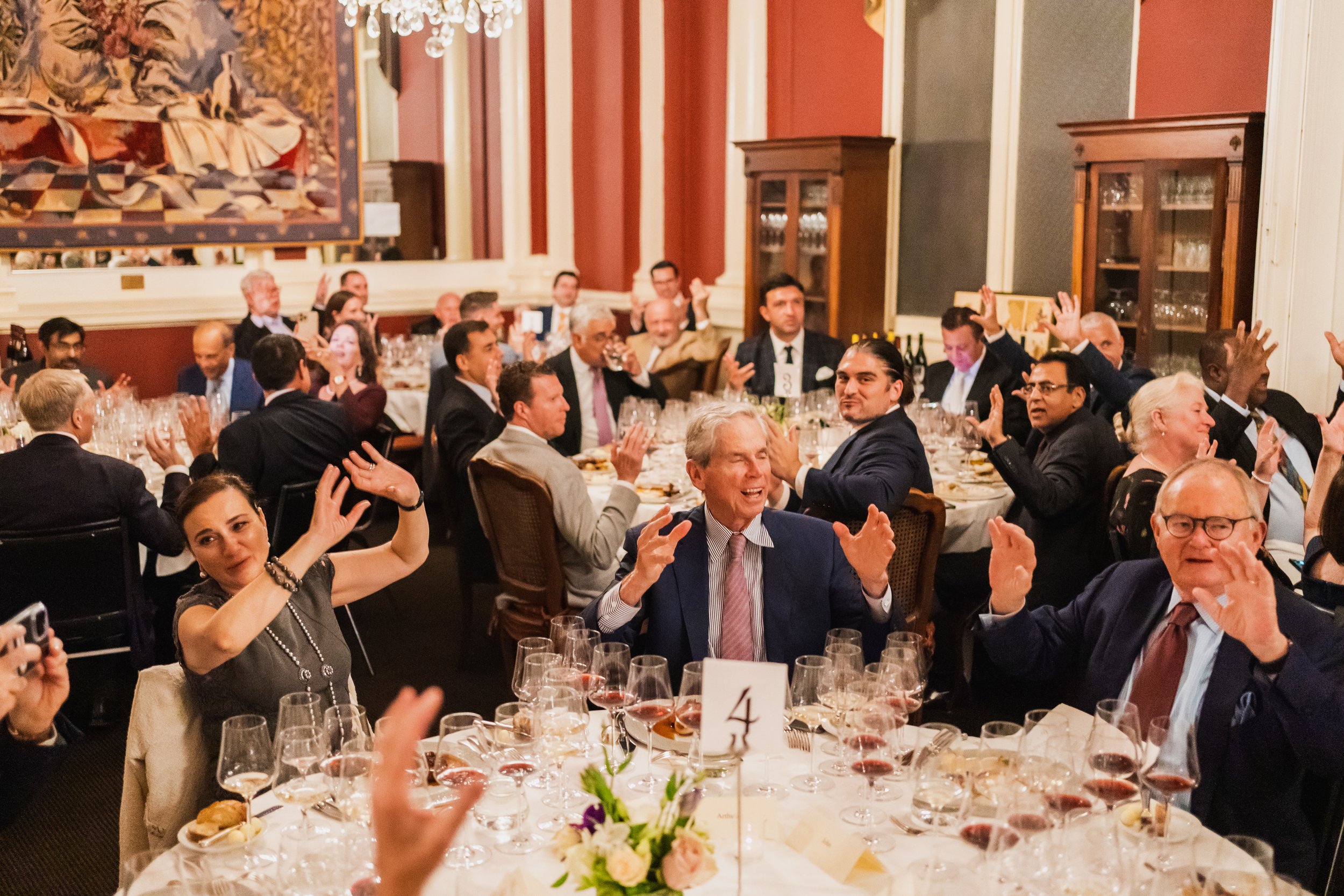 Group of people at a formal dinner celebrating, clapping and smiling in an elegant banquet room with decorated tables, glassware, and floral centerpieces.