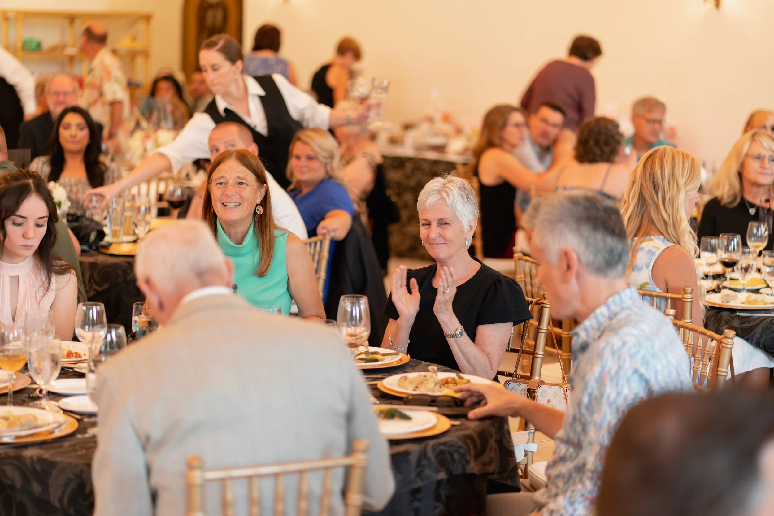 People seated at tables enjoying a meal at a banquet or celebration, with some applauding and others talking, in a well-lit room.