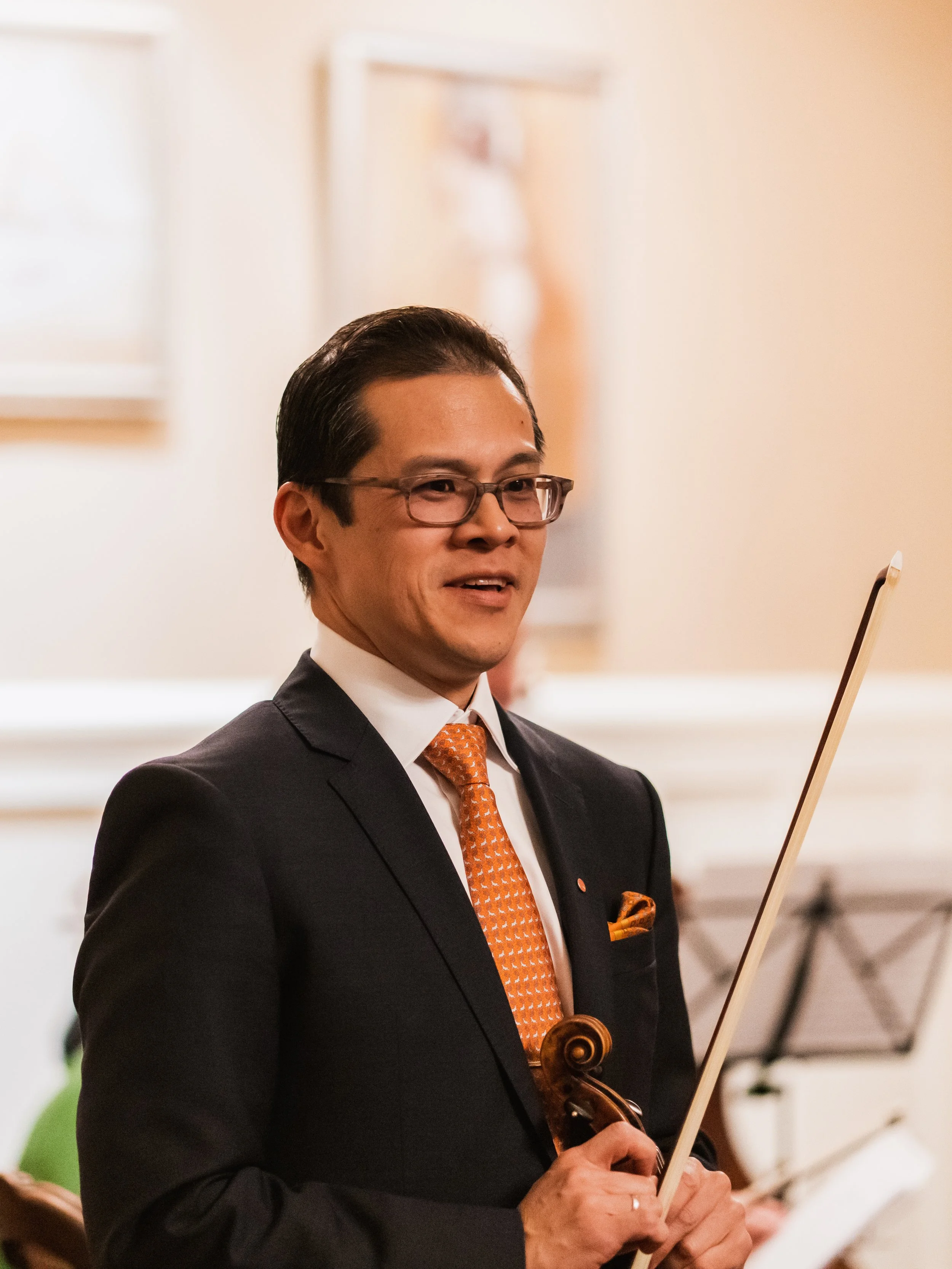 A man in a suit holding a violin and a bow, smiling, in a formal setting.