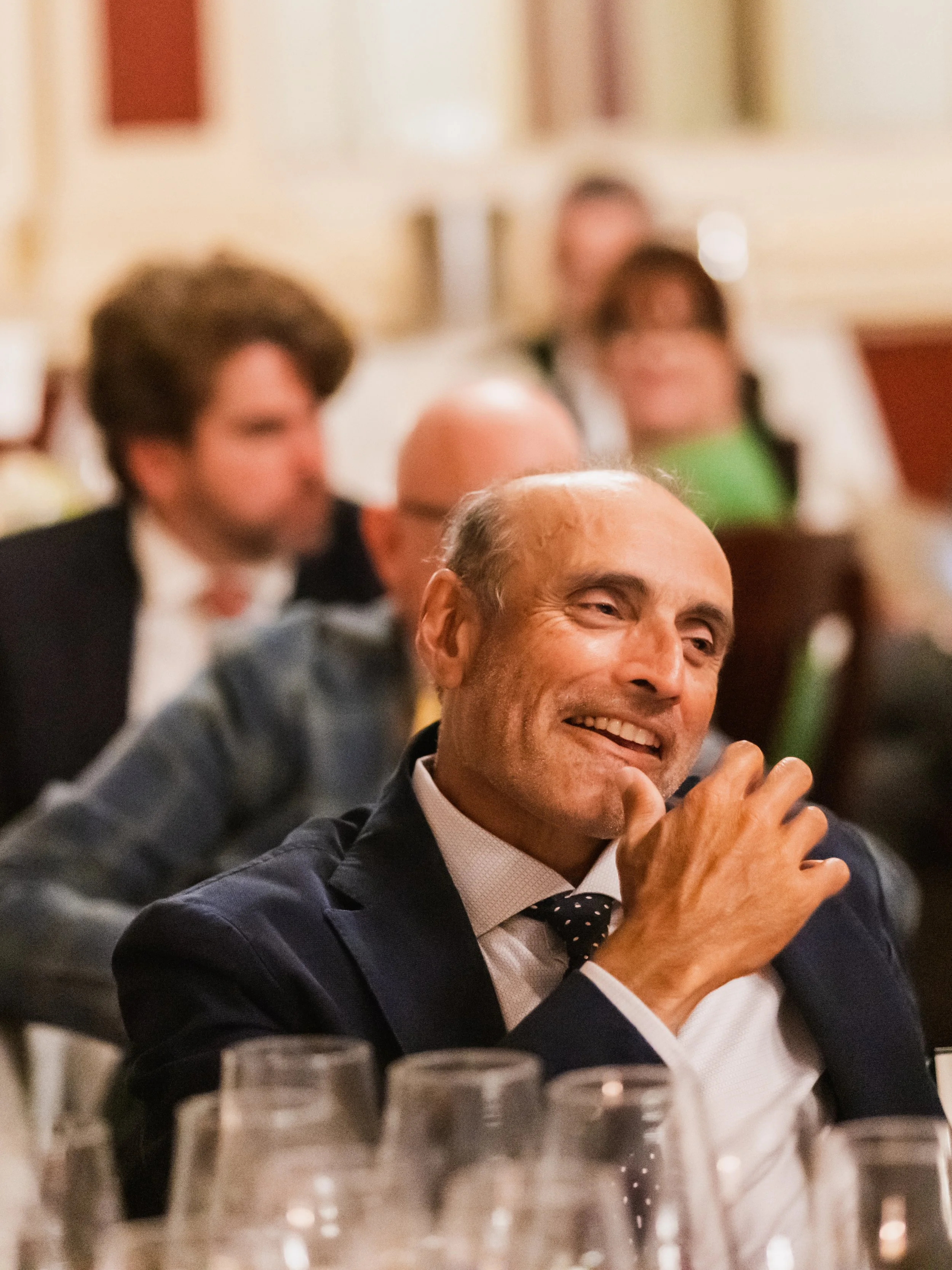 Man in formal attire smiling and laughing at a dinner event, with other guests in the background