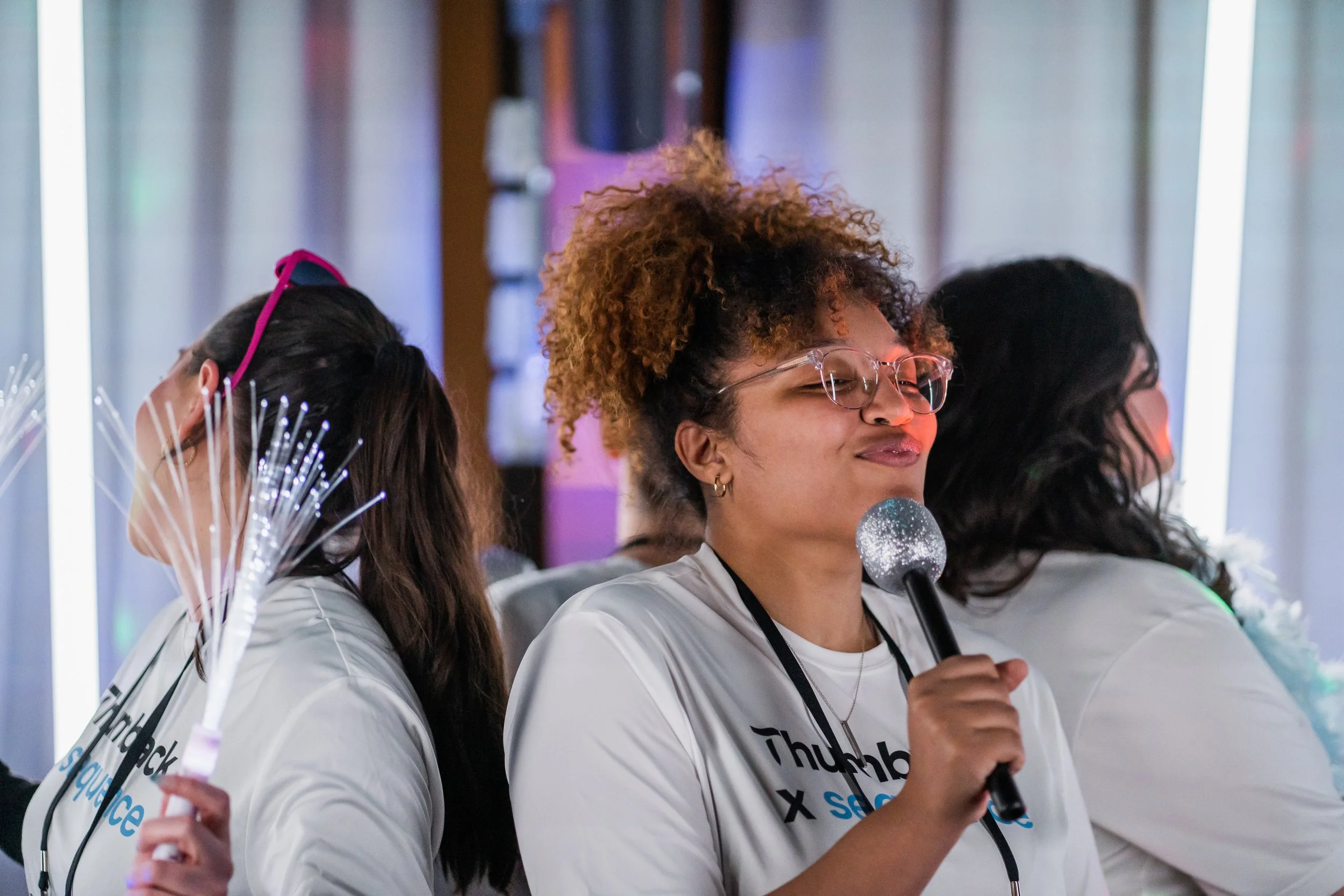 A woman with curly hair and glasses holding a microphone, with her eyes closed and sniffing it, at an event with others. The woman is wearing a white shirt with black and blue text.