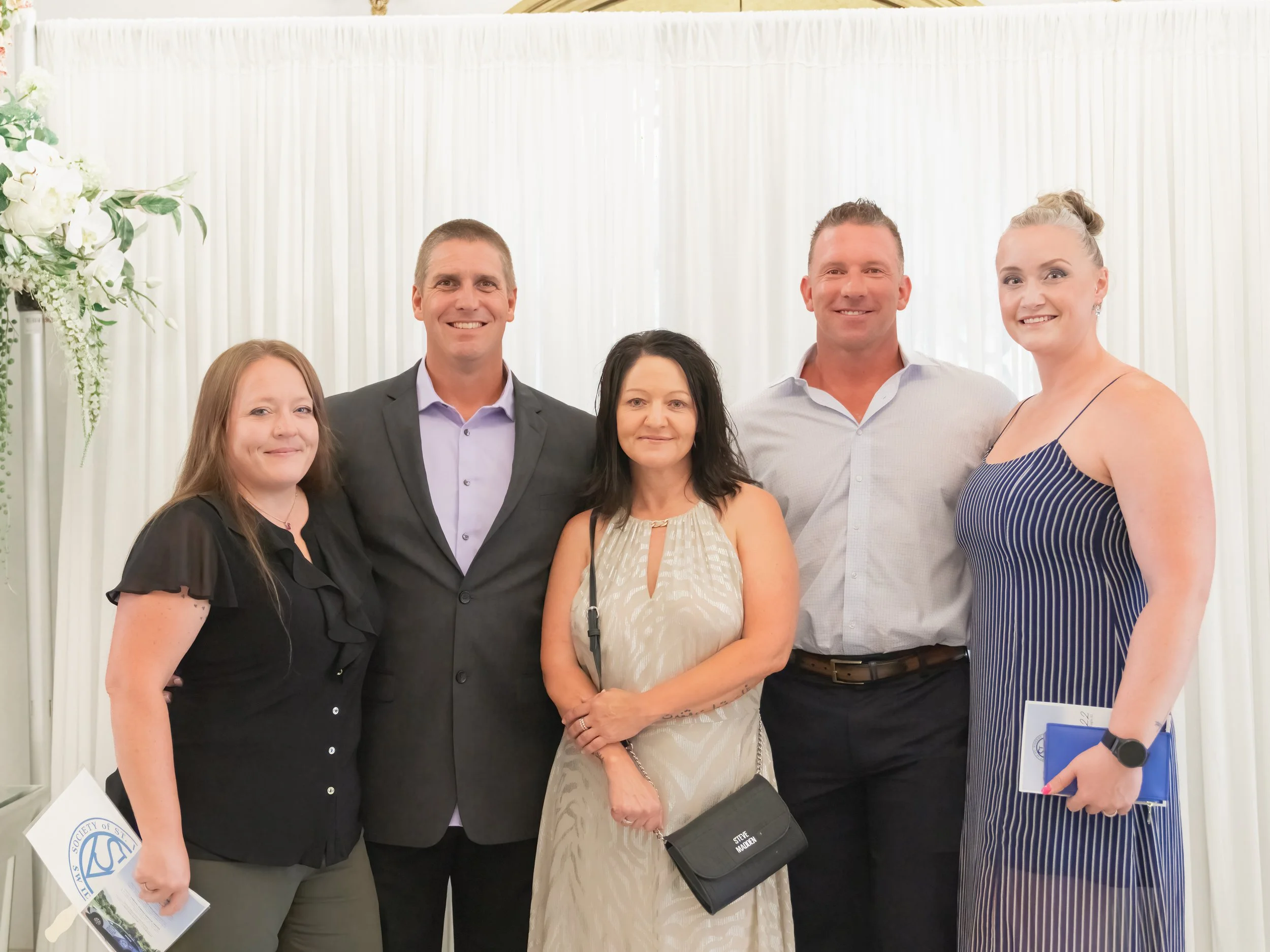 Group of six people posing for a photo in front of a white curtain backdrop at an indoor event.