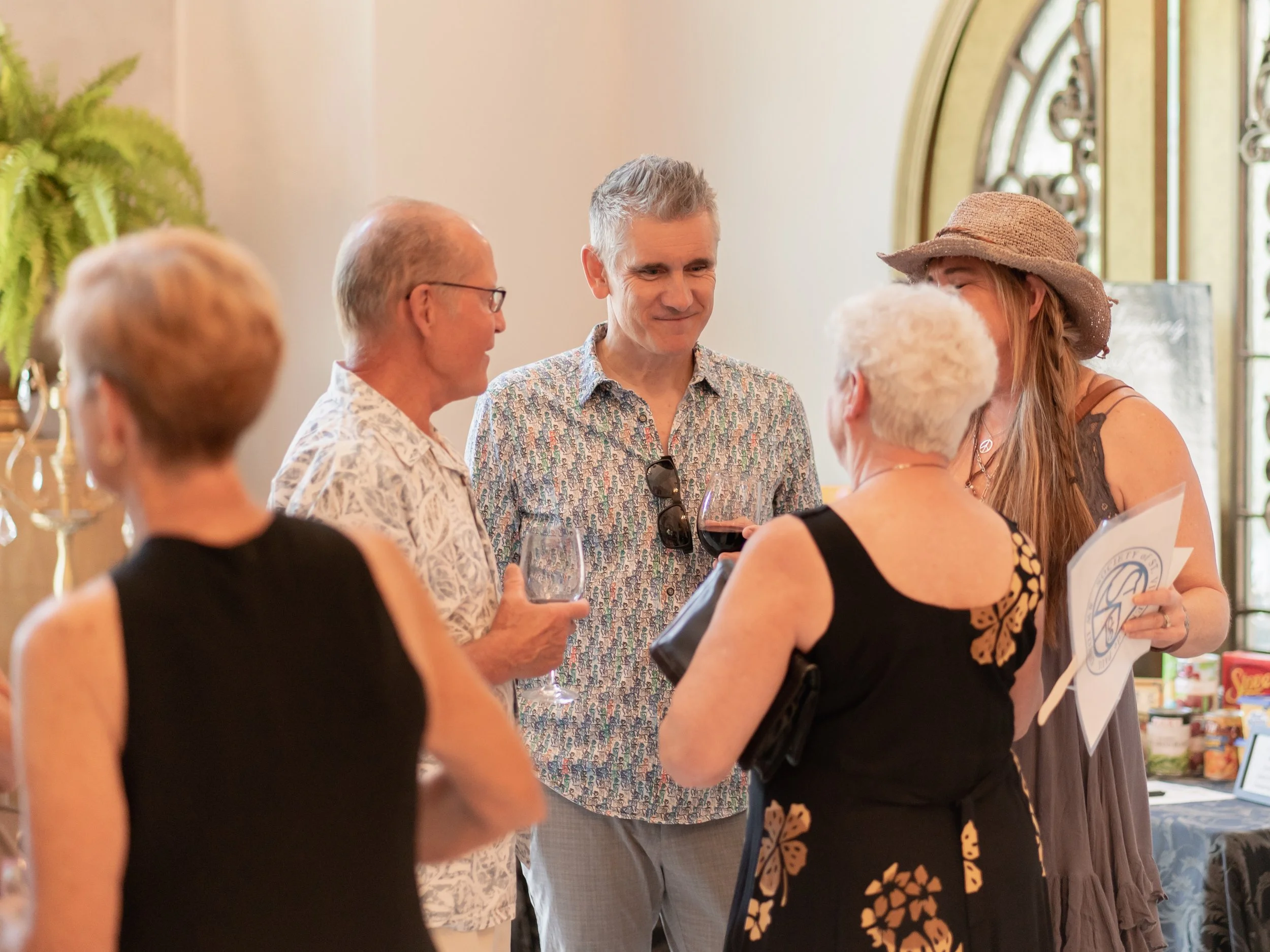 Six people standing and talking indoors, holding wine glasses, at a social gathering or party.