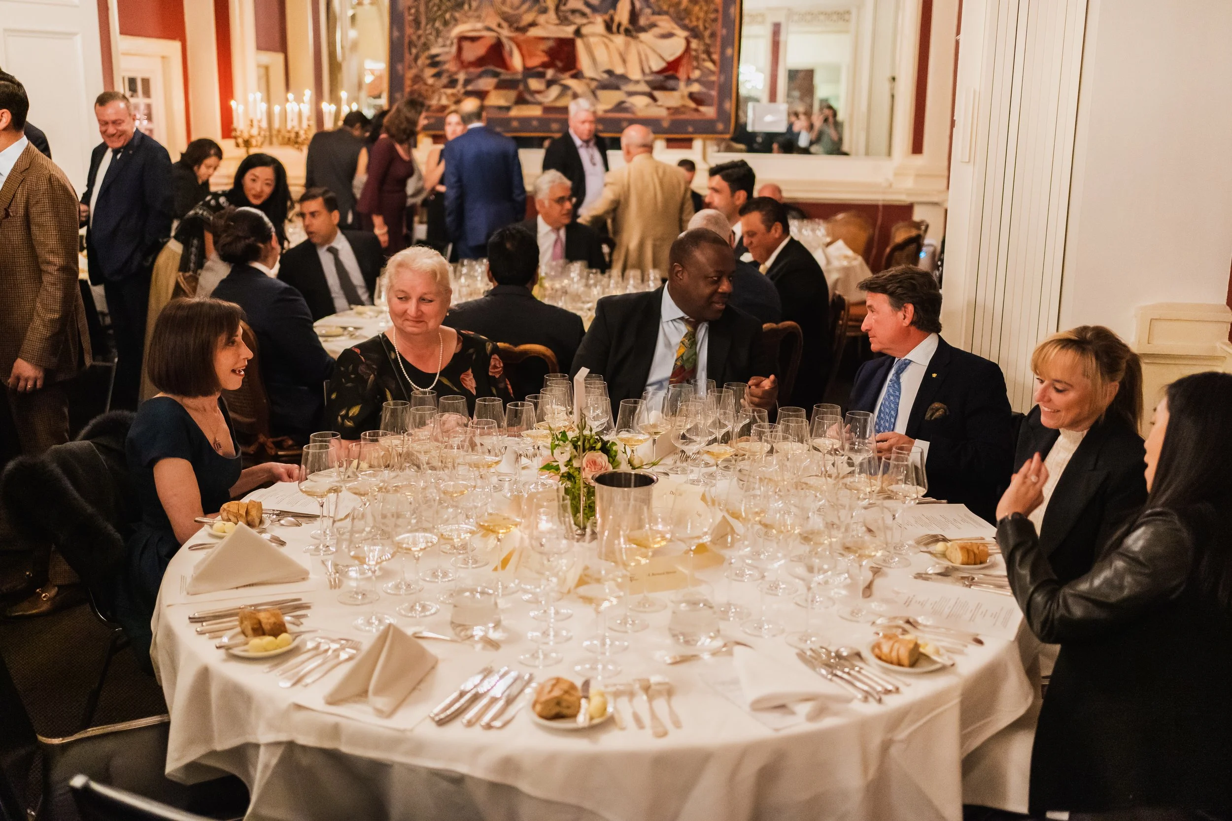 People gathered around a large round banquet table at a formal event, engaging in conversations. The table is set with white tablecloths, multiple wine glasses, plates with bread rolls, and floral centerpieces.