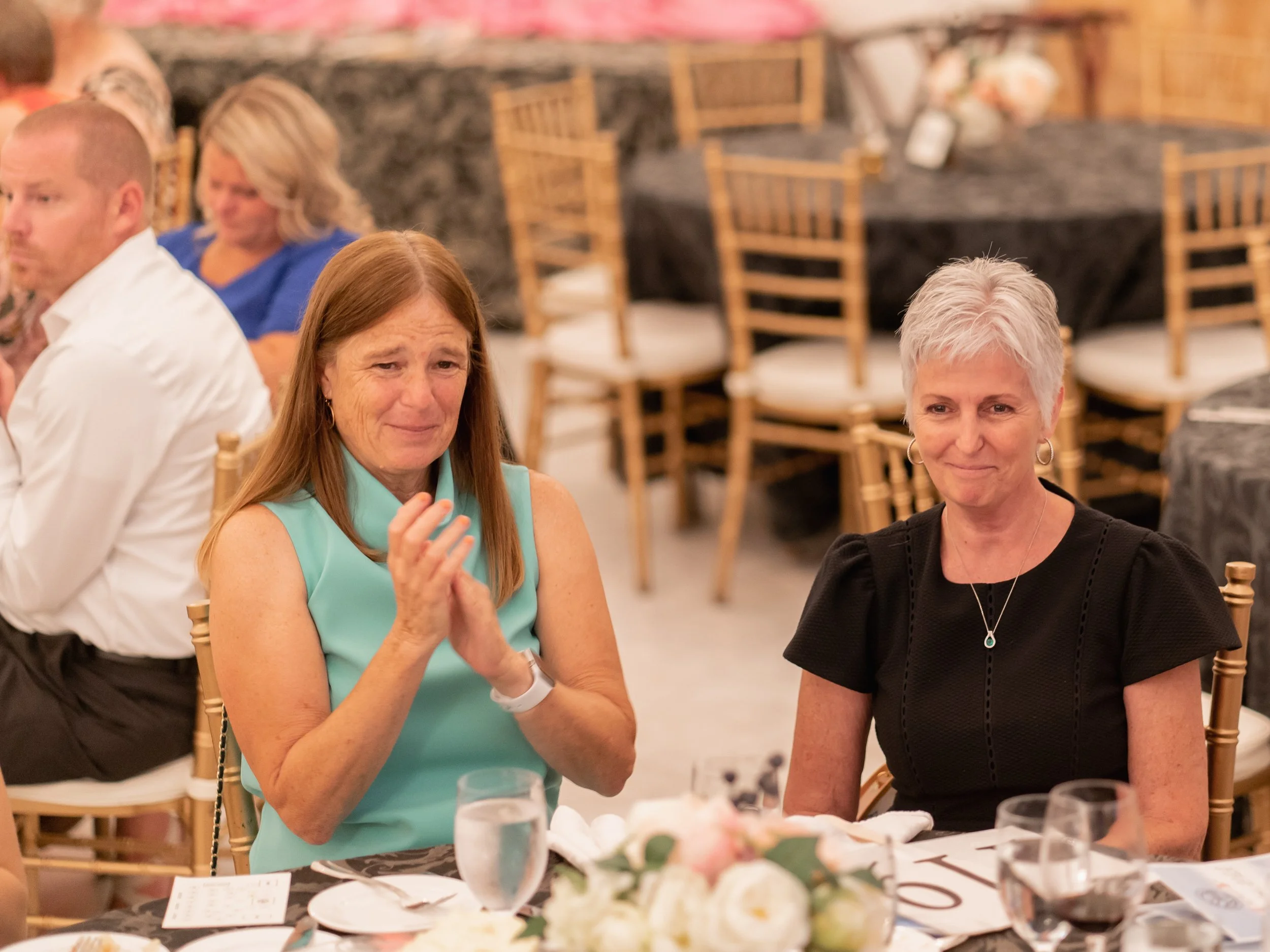 Two women seated at a table during a formal event, one with red hair wearing a sleeveless teal dress, the other with short gray hair in a black dress, with a man on the left and other guests in the background.