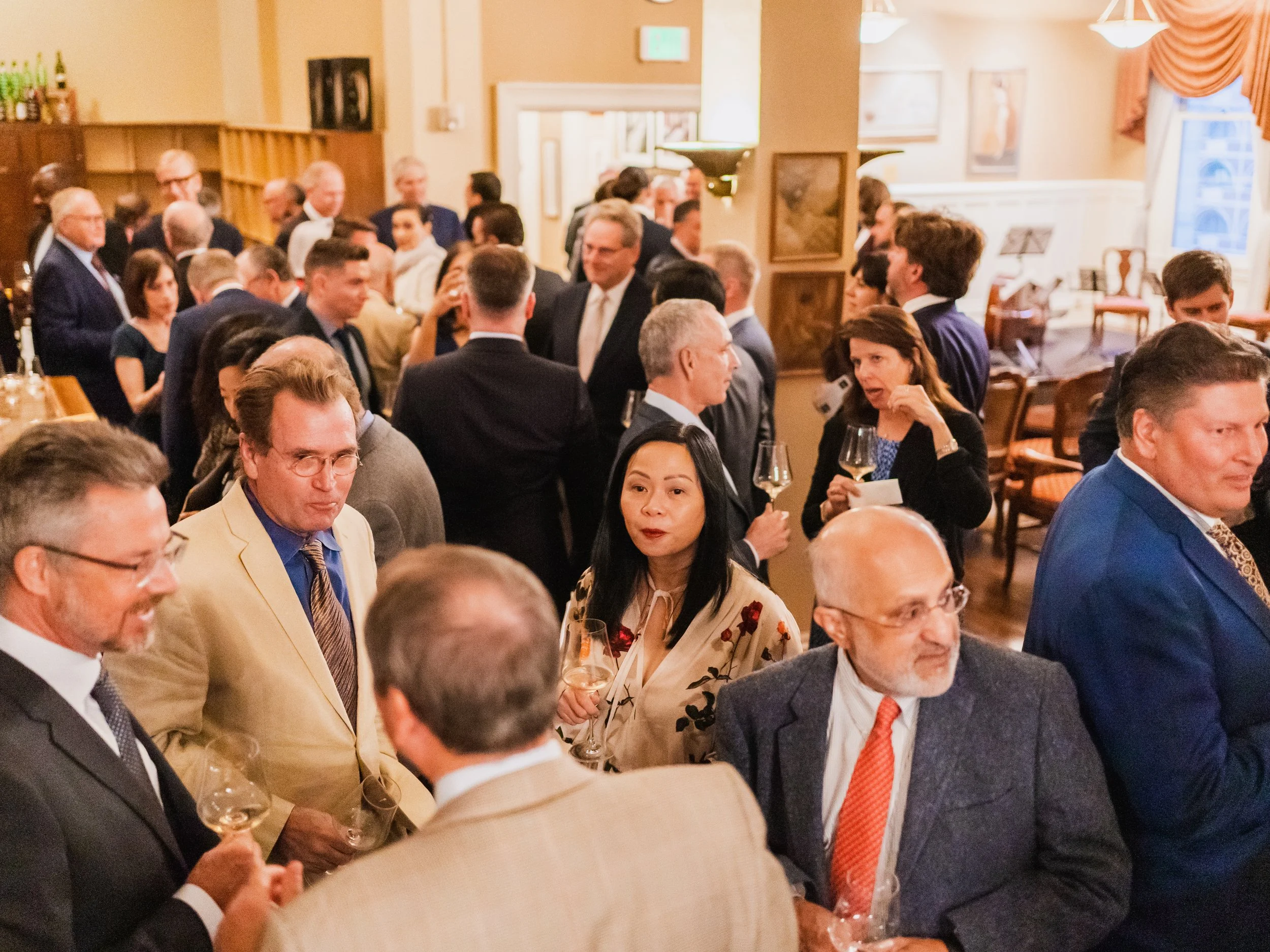 A gathering of people at a social event, holding wine glasses and engaging in conversation inside a warmly lit room.