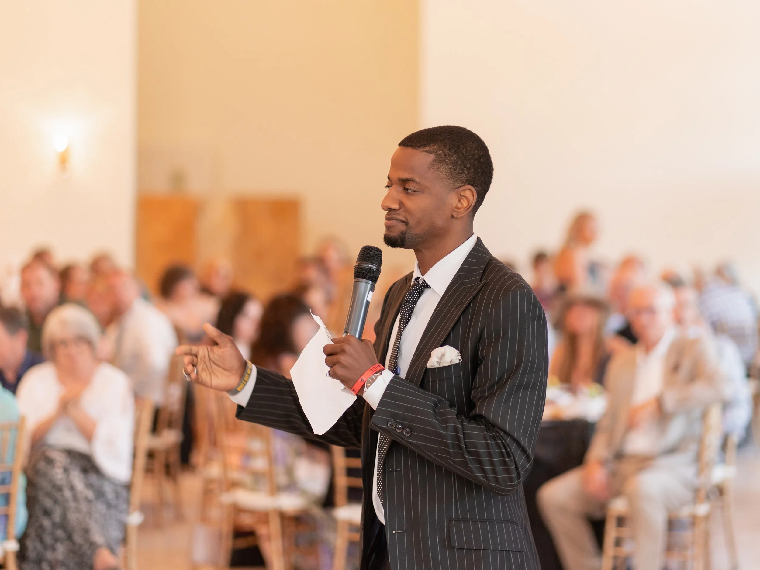 A man in a pinstripe suit holding a microphone and a piece of paper, speaking at a conference or event, with a seated audience in the background.