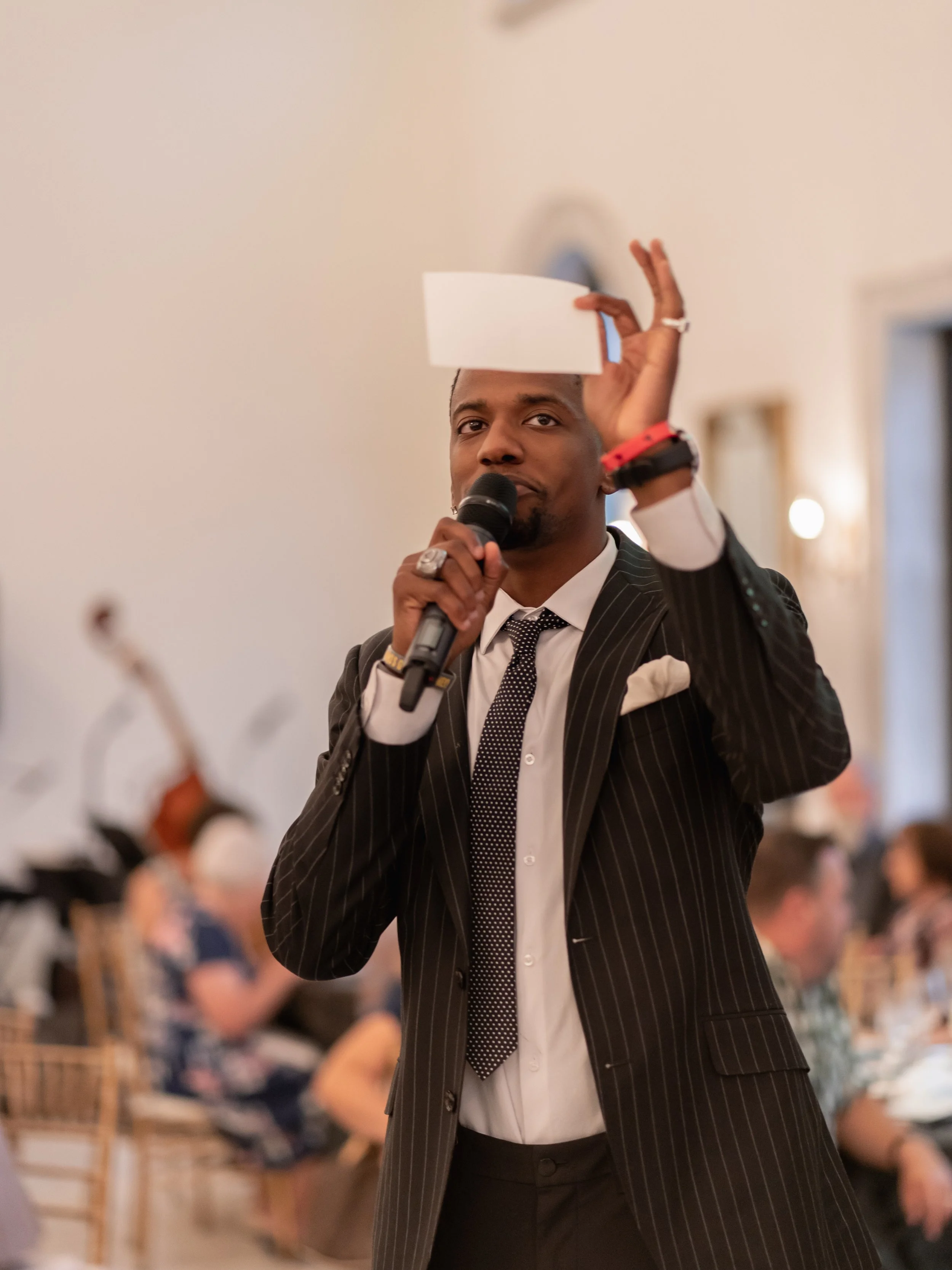 A man in a black pinstripe suit and white shirt holding a microphone and a blank piece of paper in a formal setting with guests in the background.