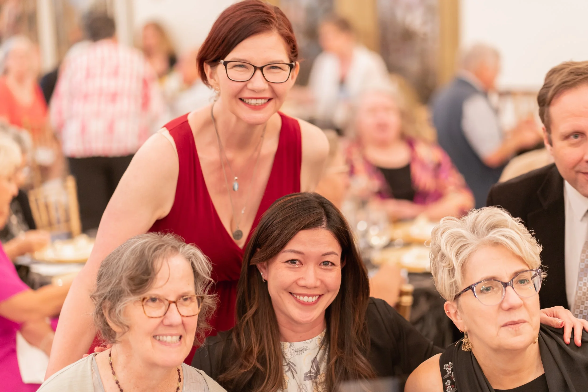 Group of women gathered at a social event, smiling for the camera, in a warmly lit room with other guests in the background.