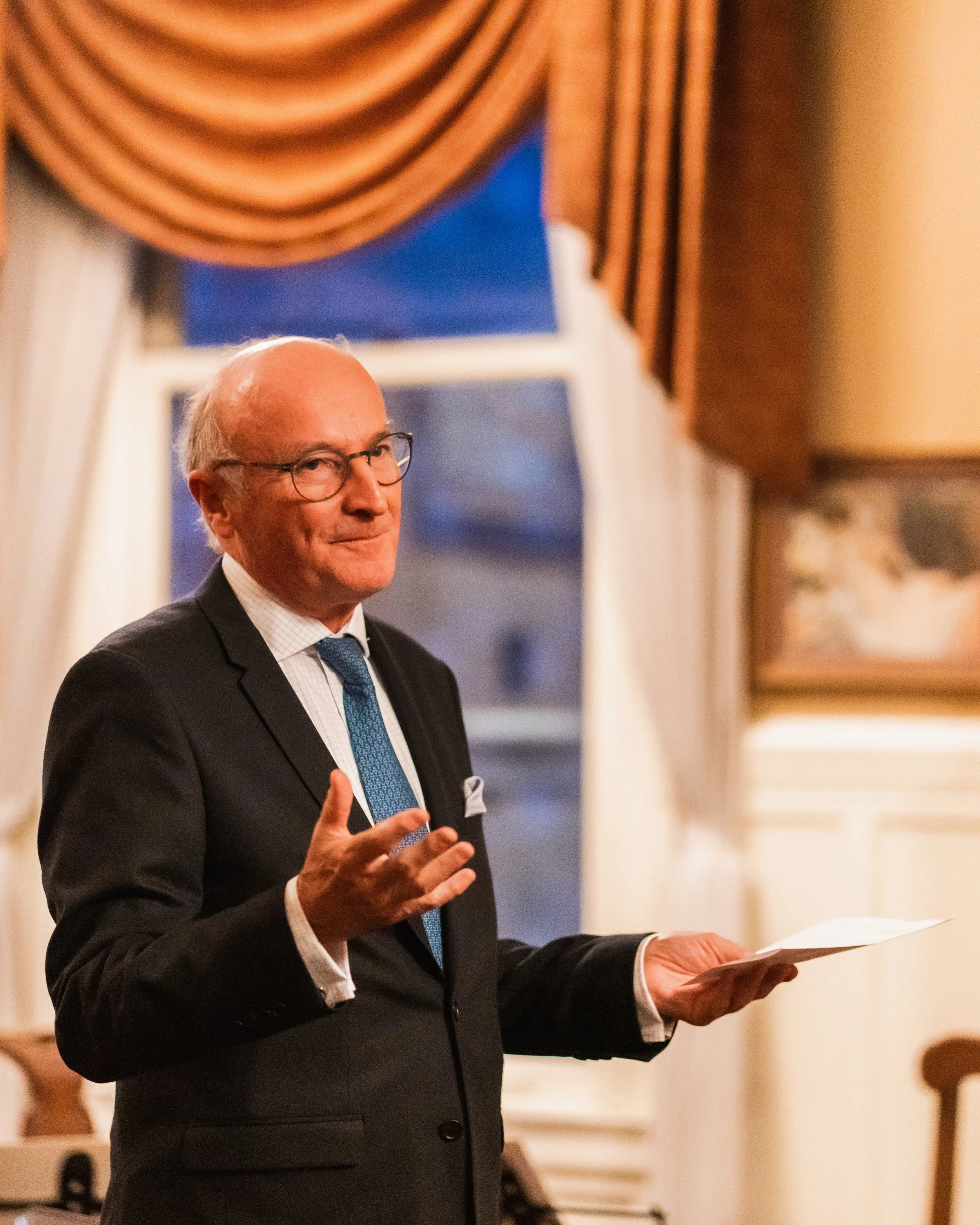 An elderly man with glasses, dressed in a black suit, white shirt, and blue tie, is speaking and gesturing with his right hand while holding a piece of paper in his left hand in a room with curtains and artwork