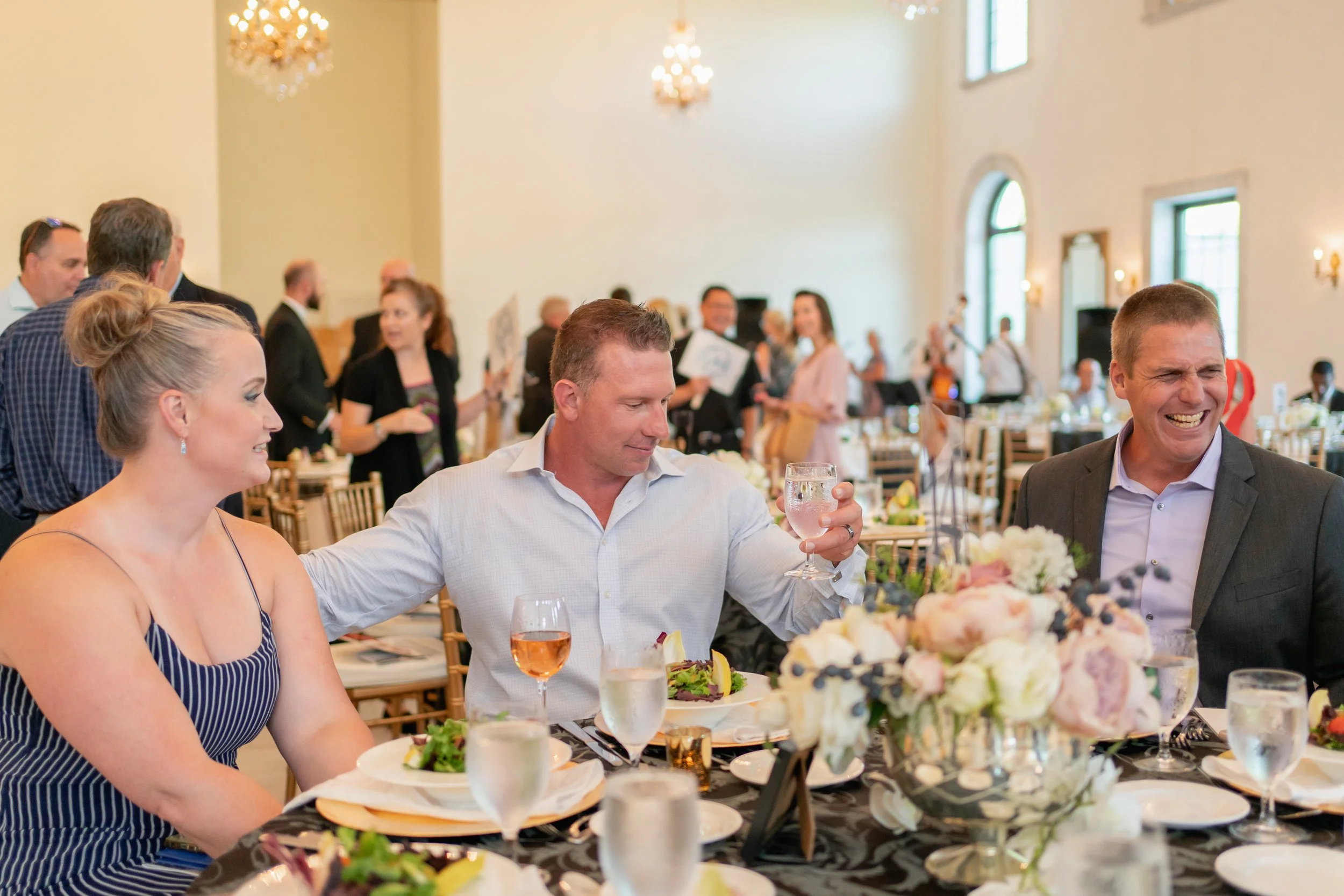 People celebrating at a wedding reception, sitting at a decorated table with floral centerpieces and enjoying drinks.