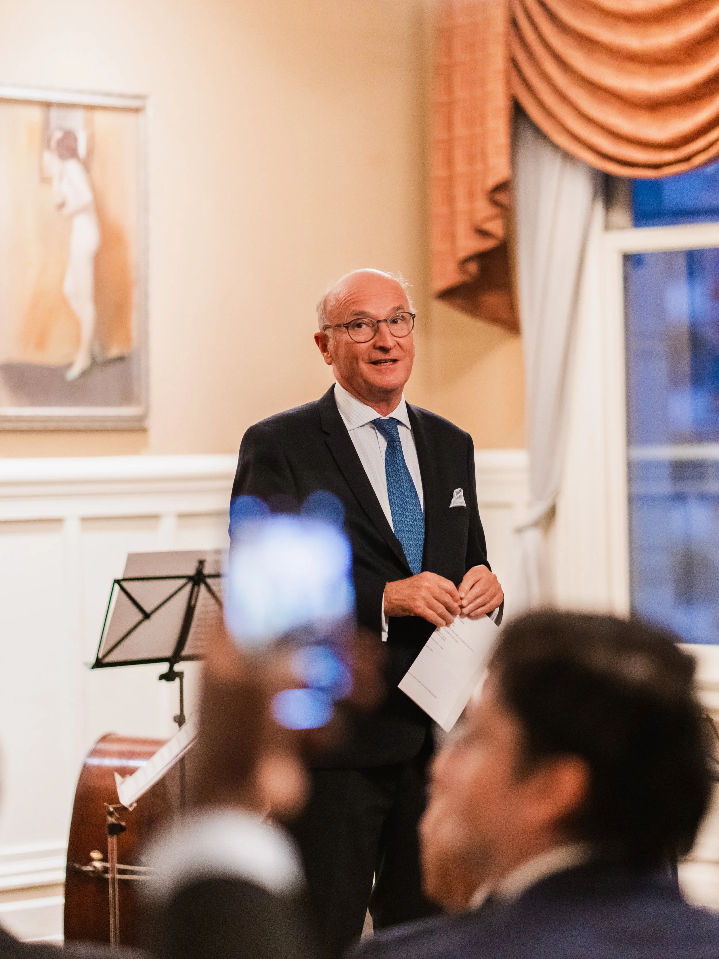 An elderly man in a suit and tie standing in a formal room, speaking to an audience, with a person in the foreground holding up a phone to record the event.