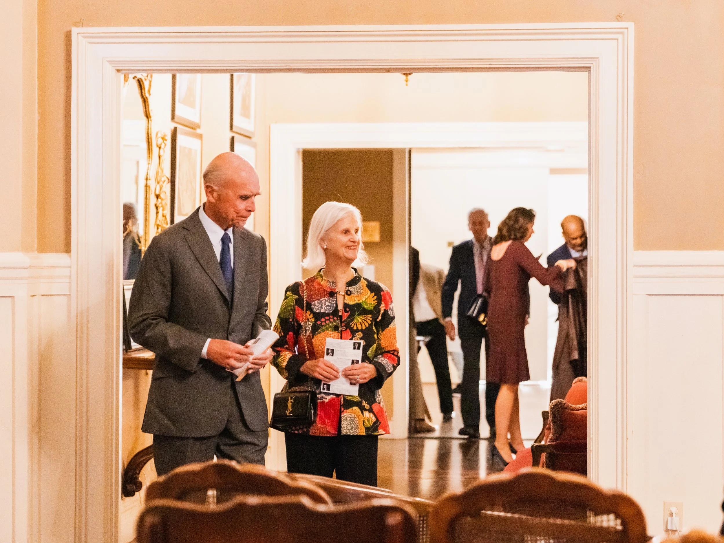 Two people, a man and a woman, standing in a room with wall decor, holding papers, and smiling. In the background, other people are mingling and talking in another room.
