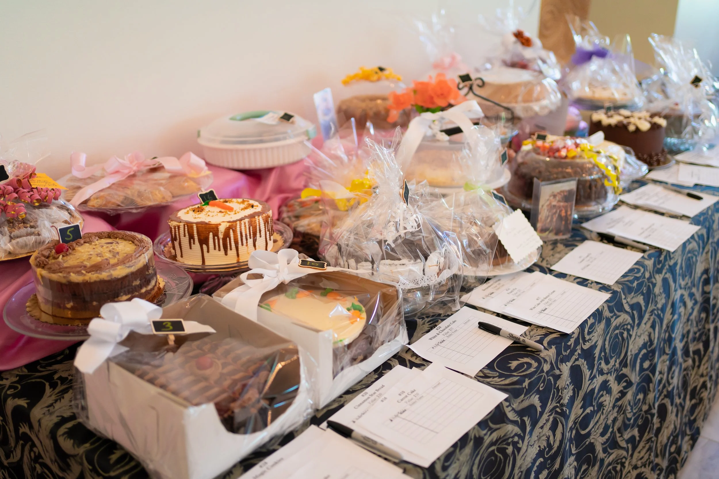 Table filled with various decorated cakes wrapped in clear plastic, some with ribbons, and notes or bidding sheets with pens underneath, at a bakery or auction event.