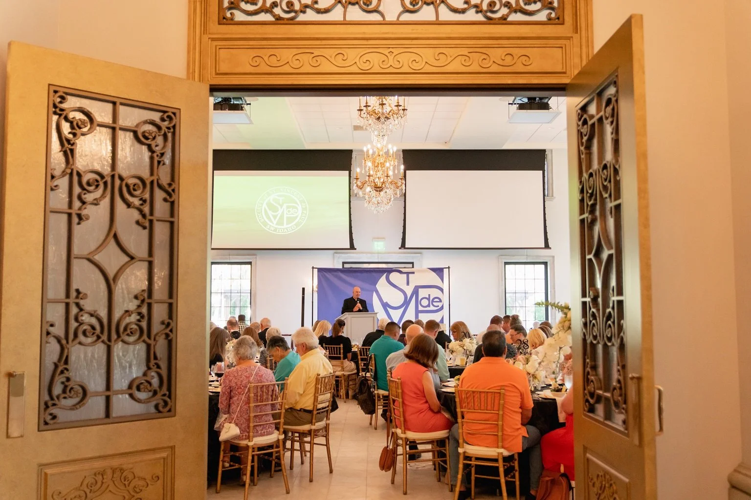 Open ornate double doors reveal a banquet hall filled with people sitting at round tables. There is a speaker at a podium on a stage with two large screens behind him, and a banner with the SVD logo. A chandelier hangs from the ceiling, and large win