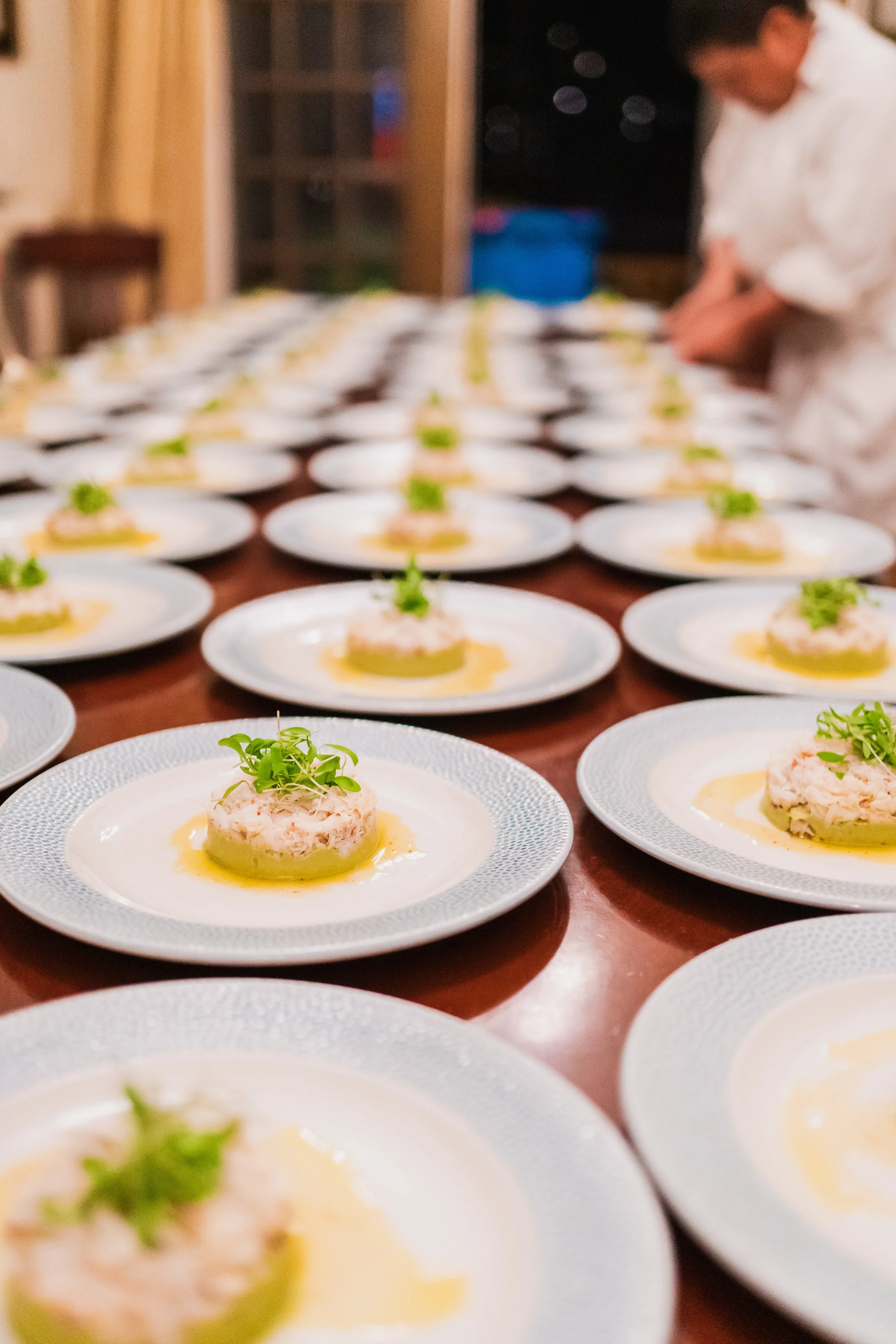 Multiple plates with a seafood dish garnished with greens on a wooden table. In the background, a chef is preparing the dishes.