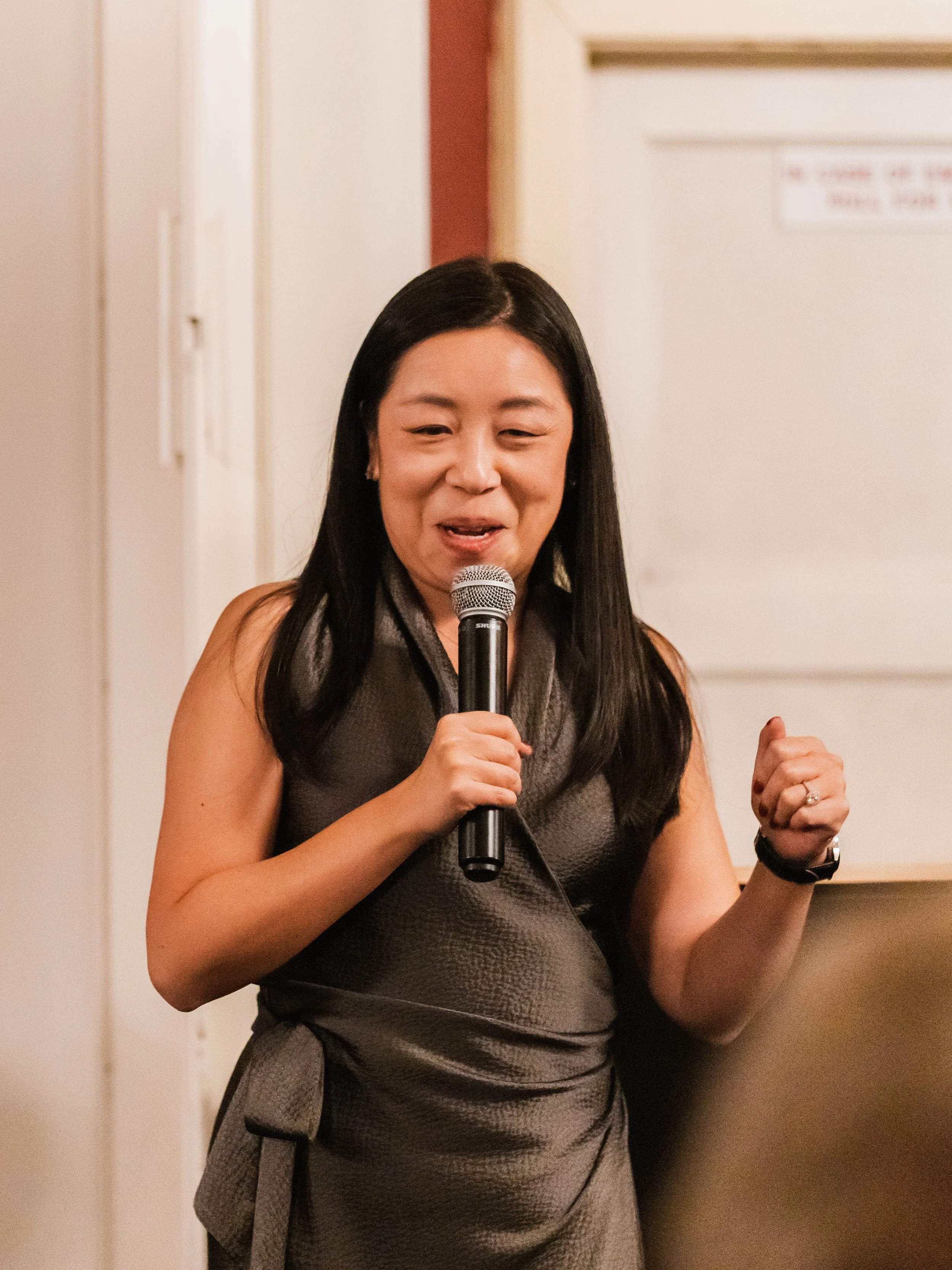 A woman with long black hair holding a microphone, speaking at an indoor event.