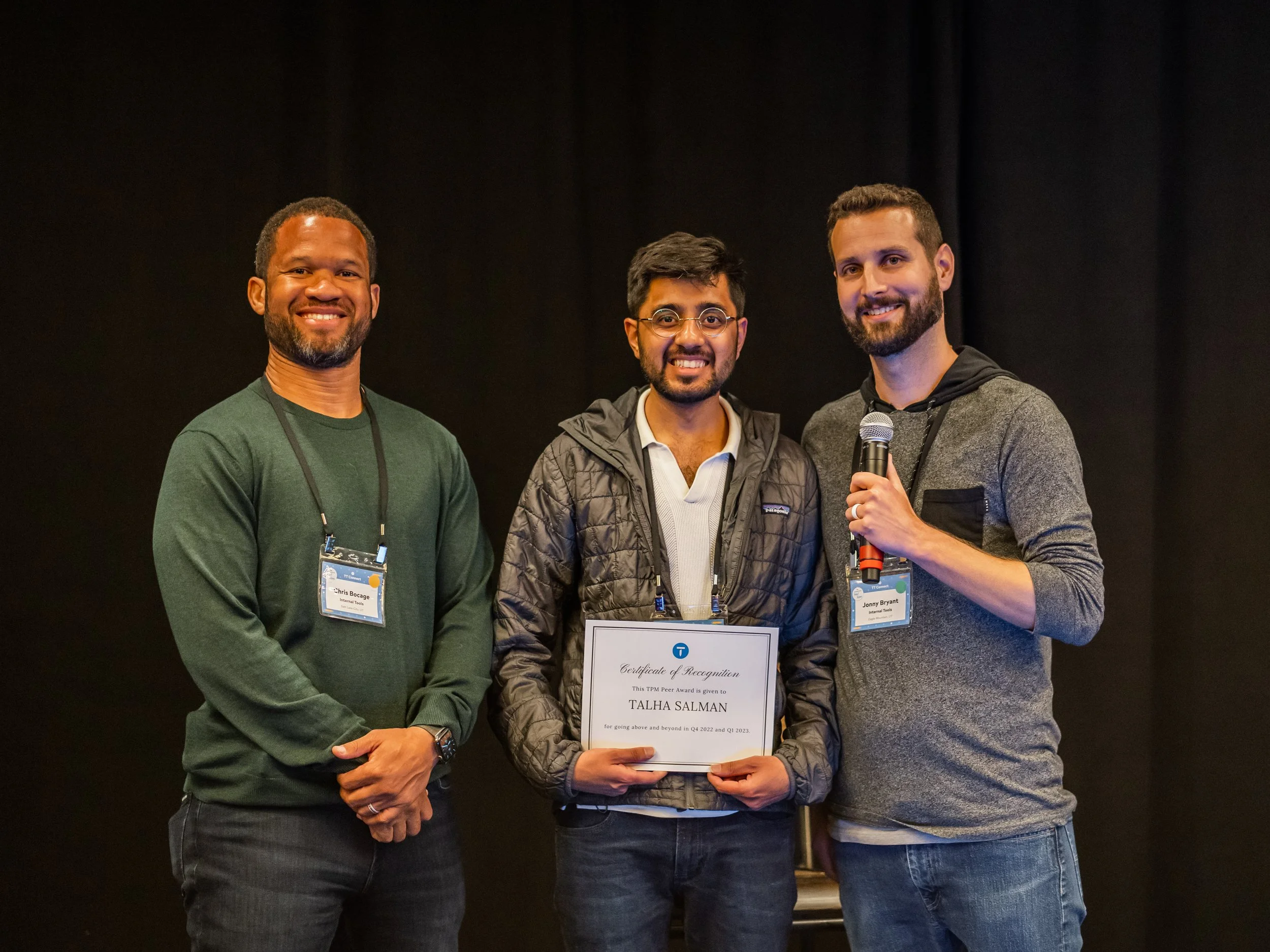Three men standing together at an award ceremony. The man in the middle is holding a certificate. The man on the right is holding a microphone. All are smiling.