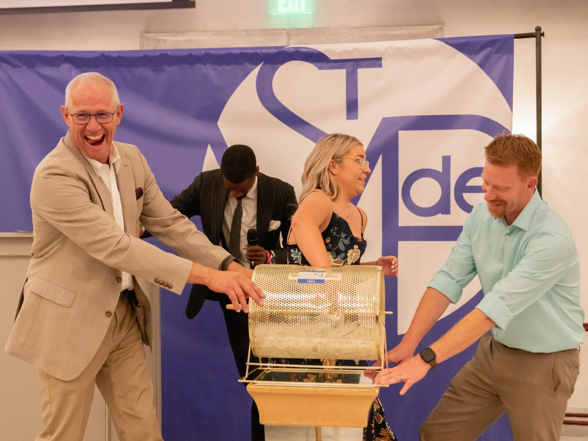 Four people, three men and one woman, participate in a lottery drawing event. They cheerfully pull tickets from a spinning drum, standing in front of a blue and white backdrop with the logo 'ST Design'.