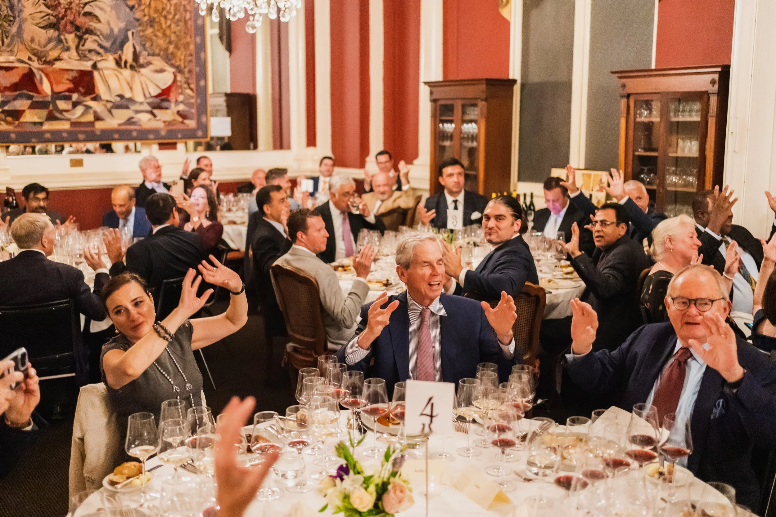 People dressed in formal attire sitting around a banquet table, many with their hands raised, at a lively dinner event in an elegant decorated room.