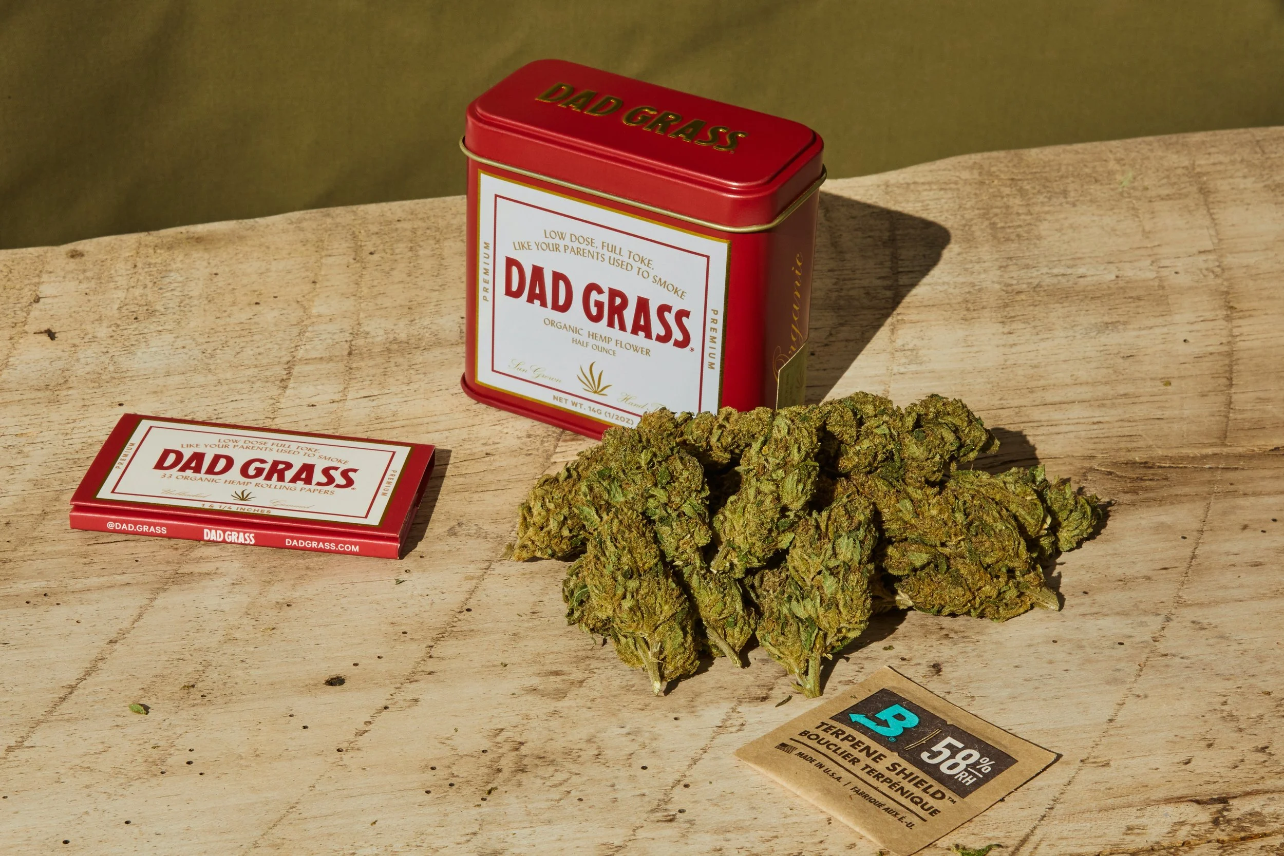 A container labeled 'Dad Grass,' a small packet, cannabis buds, and a terpene shield sticker on a wooden surface.