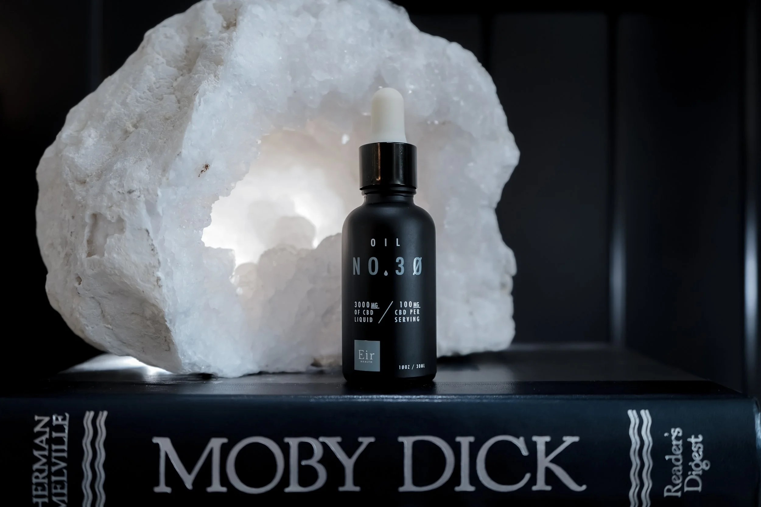 A black bottle labeled 'Oil No. 30' with a white dropper cap, placed in front of a large white salt rock, on top of a book titled 'Moby Dick' by Herman Melville.