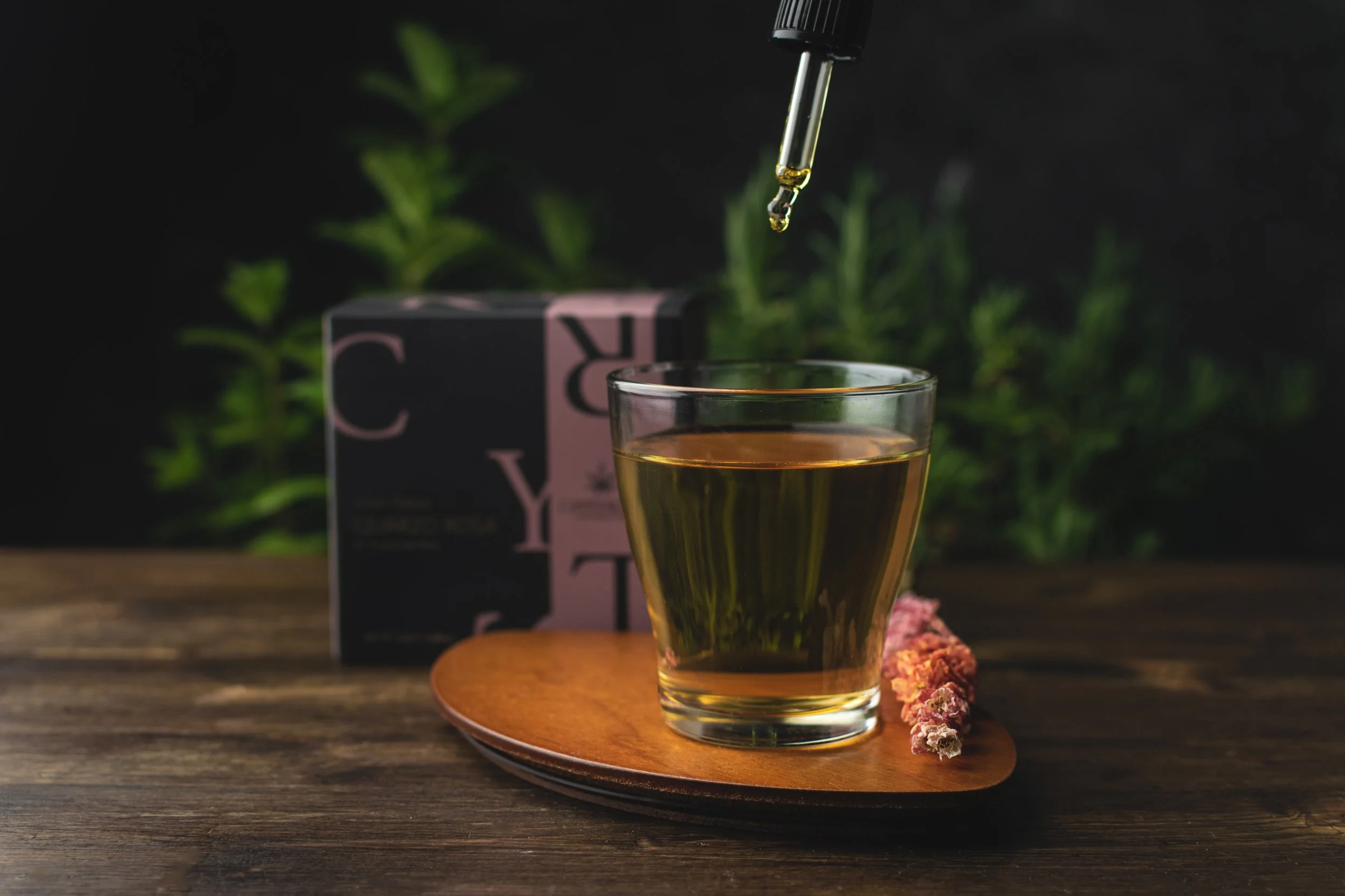 A glass of herbal herbal tea on a wooden coaster, with a pipette dropping oil into it, a box labeled 'C', and green herbs in the background.