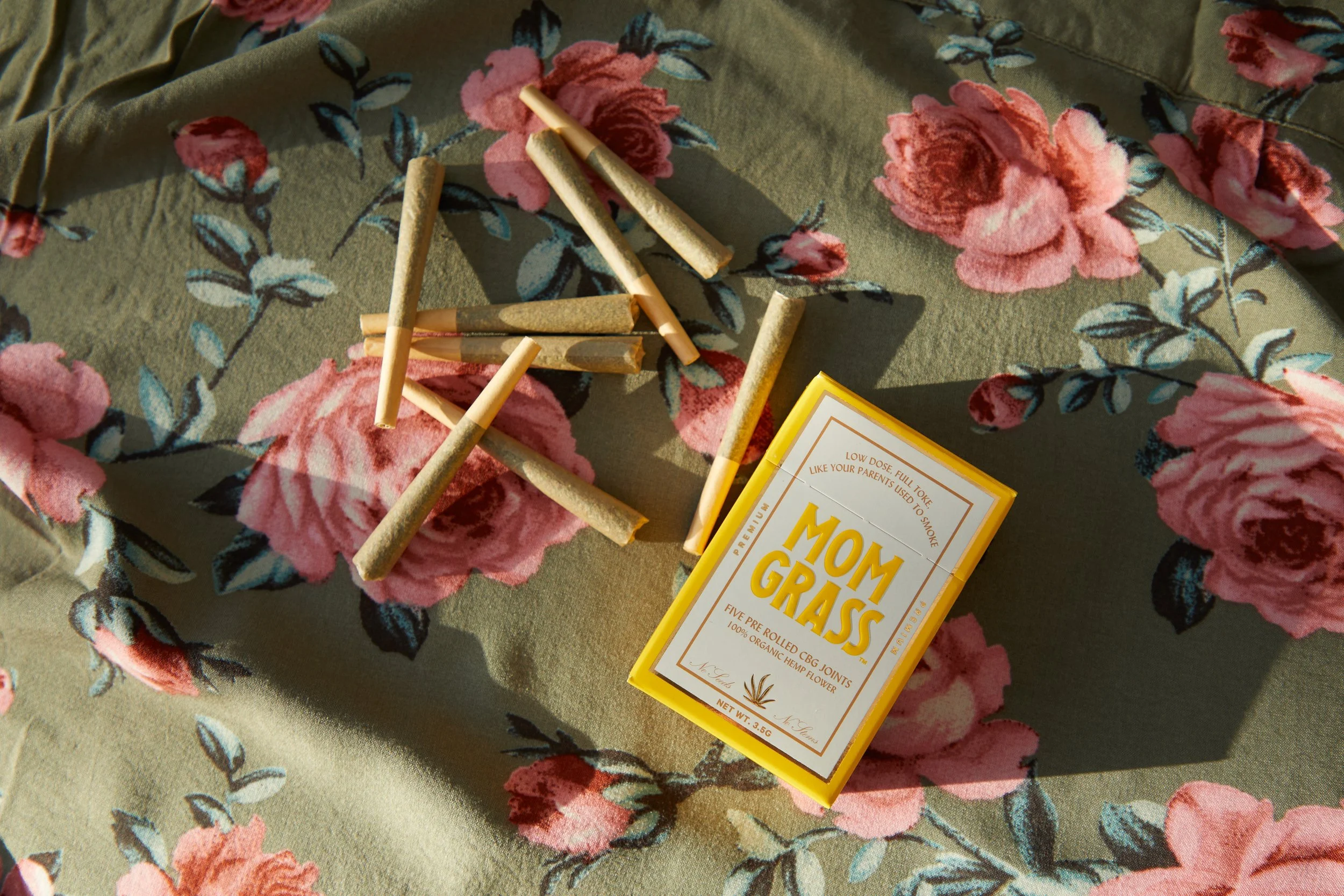 A box of Mom Grass CBD joints and several loose joints scattered on floral-patterned fabric.