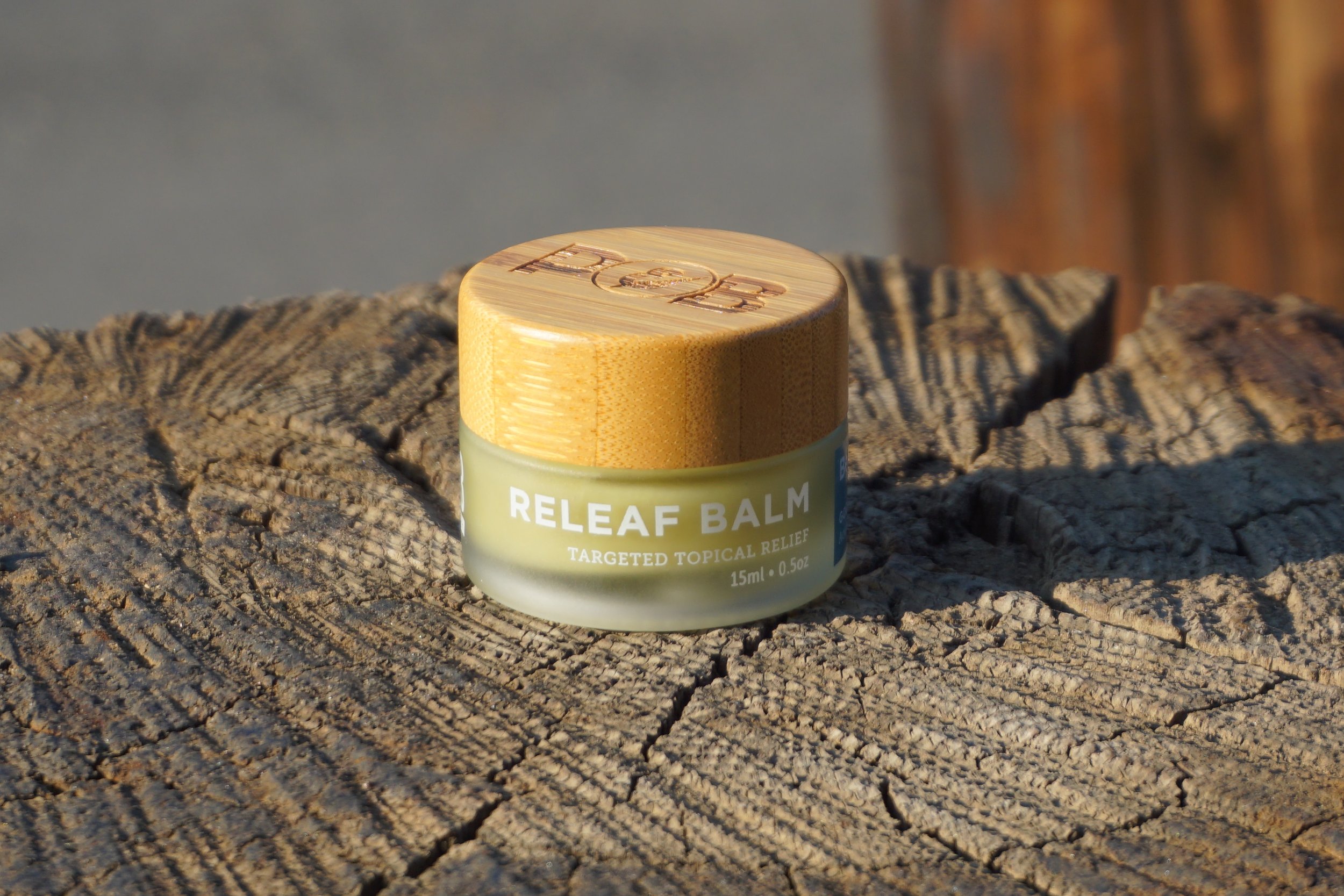 A small jar of RELIEF BALM with a wooden lid, placed on a textured wooden surface outdoors, with natural lighting.