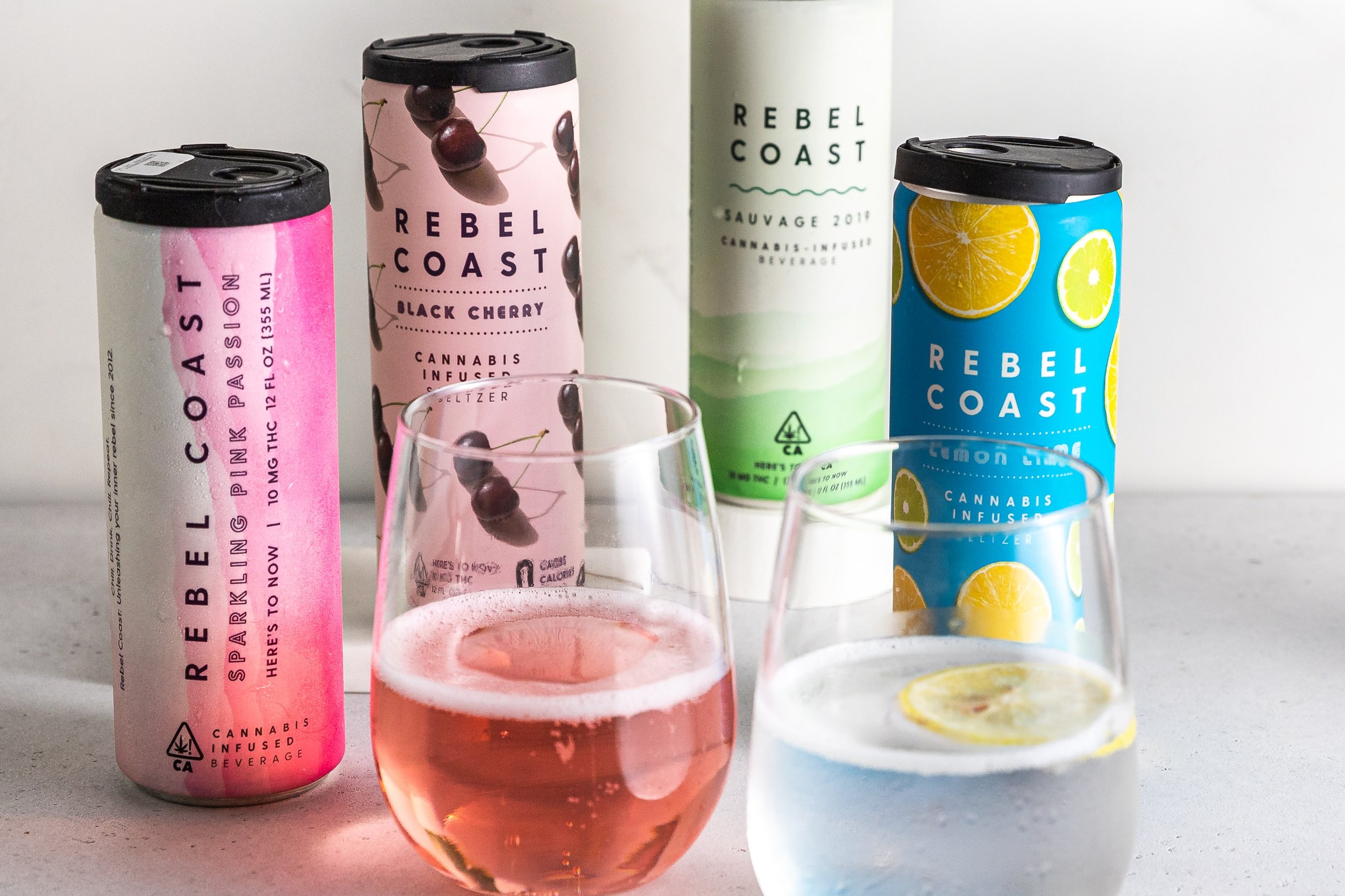 Four cans of Rebel Coast cannabis-infused beverages arranged on a white surface, with two glasses filled with colorful drinks in front. The cans are pink, black cherry; green, Sauvage; blue, lemon citrus; and light green, unknown flavor.